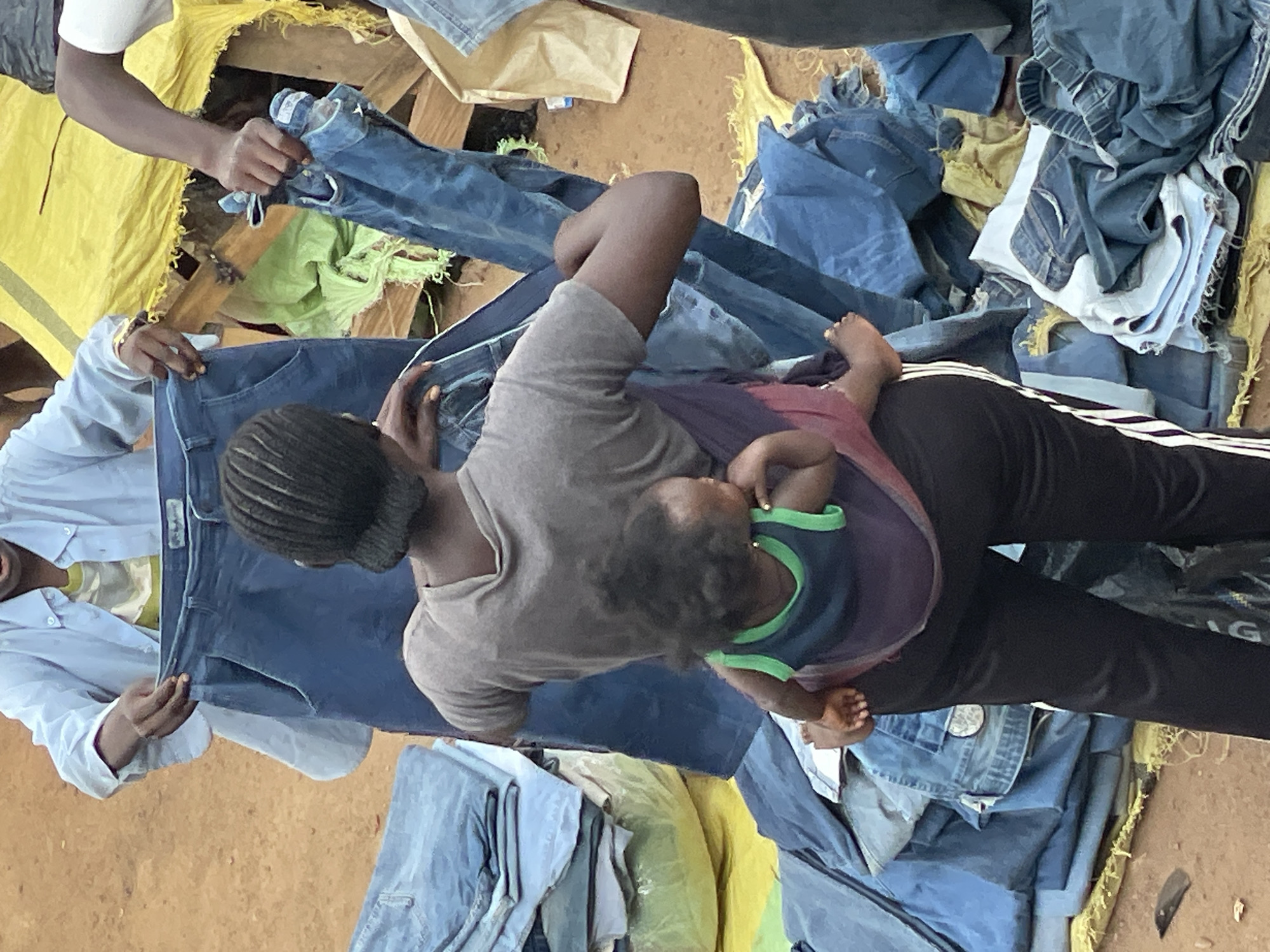 Captured at an undisclosed location within the city of Unknown, Unknown, the image presents a complex scene focused around the sale of secondhand clothing. A woman, her hair styled in short braids, is centrally positioned, carrying a small child in a sling across her chest. The child's small hand is visible, resting against the woman's body. The woman is situated amidst a pile of denim jeans and other clothing items, indicating a marketplace or informal trading environment. Several individuals are actively engaged in sifting through the clothes, suggesting a transaction or bargaining process. The setting includes elements such as yellow tarps and what appears to be simple wooden structures in the background, which could be part of a makeshift stall or display. The overall composition of the scene points to a busy, possibly crowded, location with a focus on the exchange of goods.