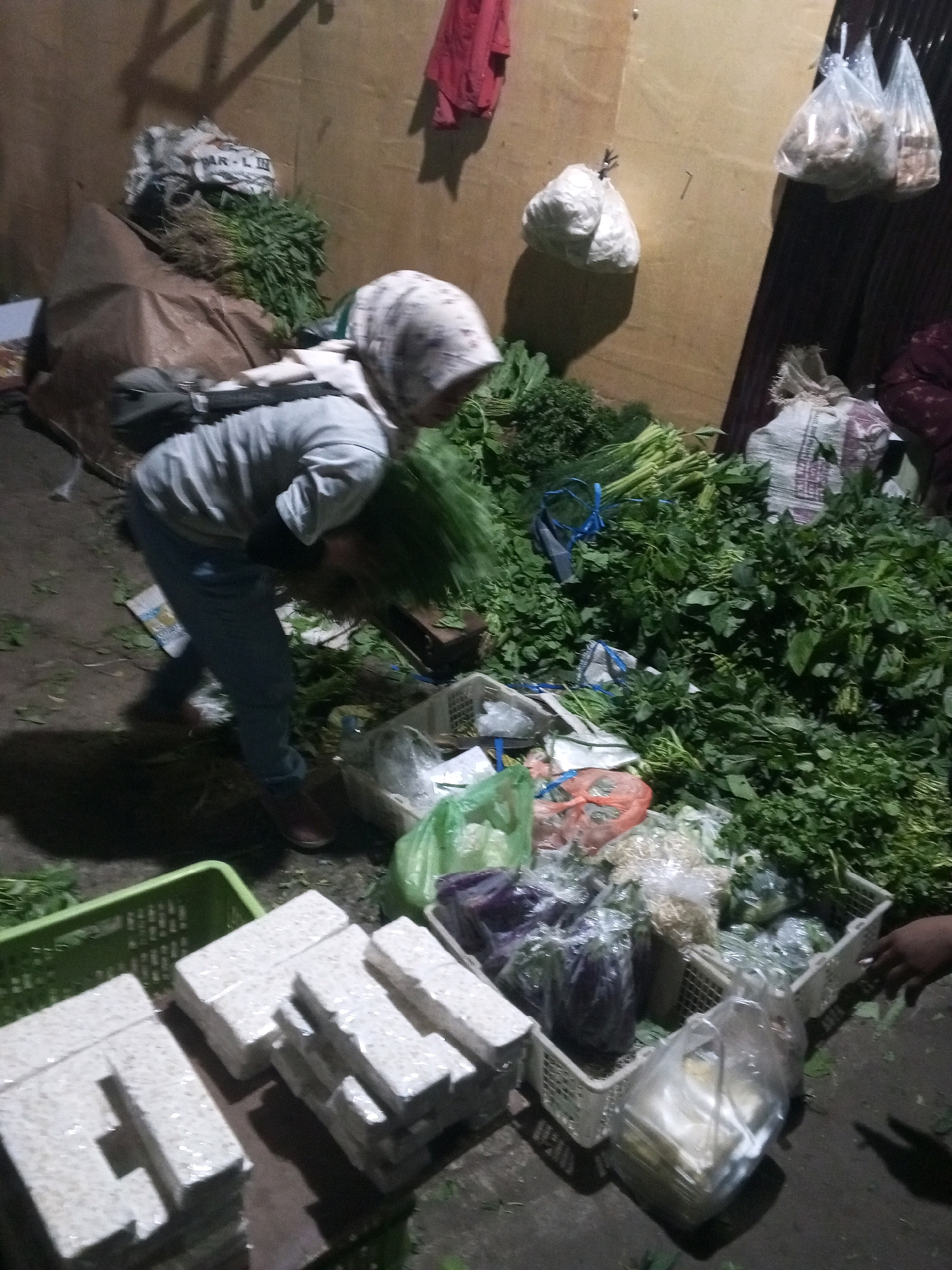 A market stall is depicted, likely in Gentan, Indonesia, with a person wearing a light-colored headscarf and grey shirt bent over a large pile of green leafy vegetables. The person is also wearing blue jeans and dark shoes. Several other baskets and bags filled with produce are visible around the stall. In the foreground, stacks of rectangular, individually wrapped items, possibly tempeh, are arranged on a surface. The environment appears to be an outdoor or covered market setting, with a dirt or concrete ground and a rough wall in the background. Bags of produce hang from the wall. The lighting is dim, suggesting it might be early morning or evening.