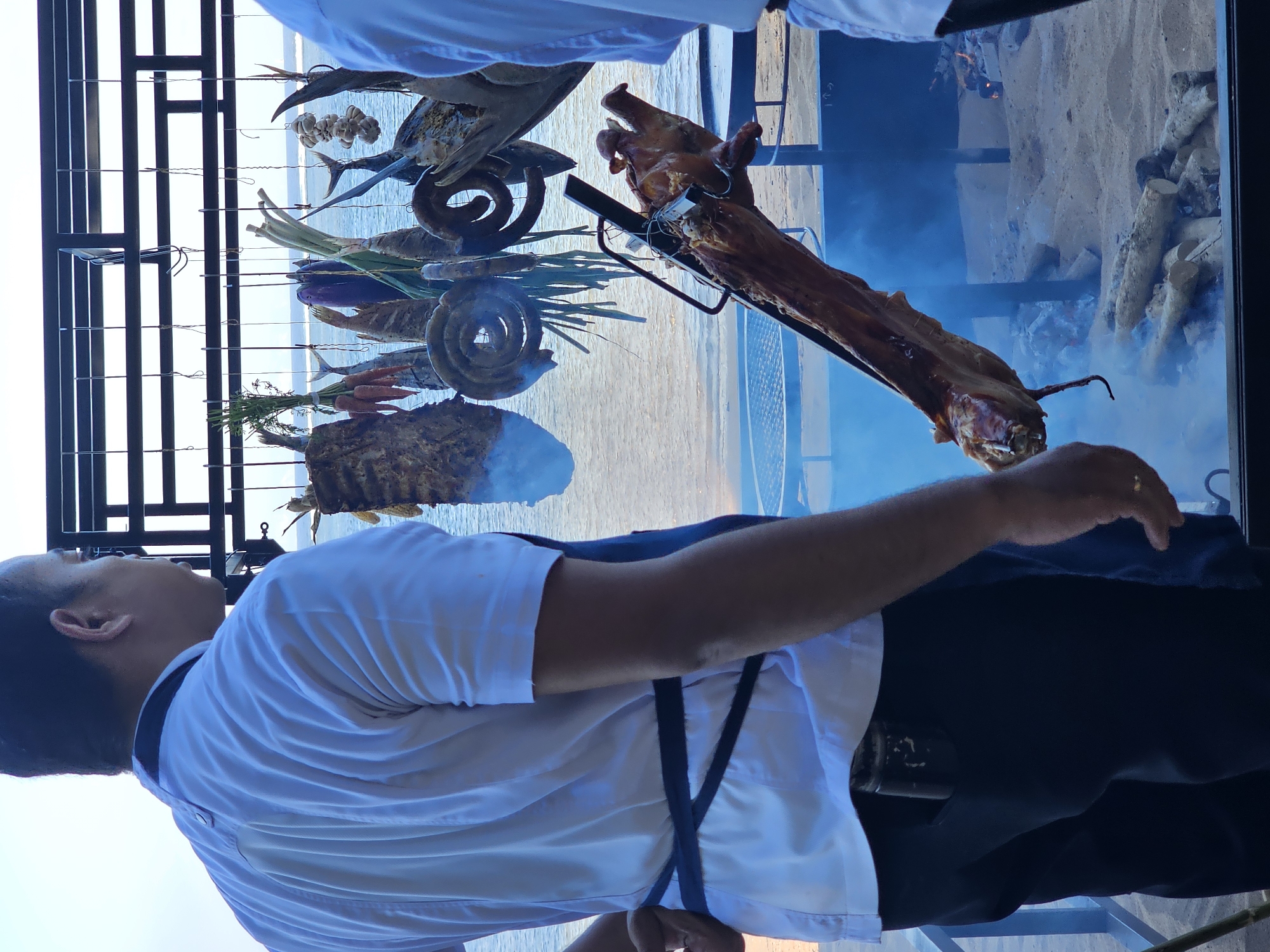 The image captures a scene of an outdoor culinary event, likely a barbecue or food preparation area, set against a backdrop of the ocean. In the foreground, a person wearing a white chef's uniform with a dark apron is partially visible from behind. They are positioned to the left of the frame, facing towards the right.
The central focus is a whole pig being roasted on a spit over a fire. The pig is positioned at an angle, its head and front limbs clearly visible, appearing browned and cooked. Smoke rises from the fire pit, which is filled with logs and embers, contributing to the atmosphere of cooking.
Above and behind the roasting pig, a collection of various foods are suspended from a metal rack. These include what appear to be dried fish, sausages, vegetables like carrots and leeks, and possibly some root vegetables or other prepared items. The sea and the horizon are visible in the distance, suggesting a coastal location. The ground appears to be sandy.
The overall impression is one of a rustic, open-air food preparation setting, likely associated with a restaurant, resort, or special event. Given the context of outdoor culinary events and potential tourism, such a scene could be found in areas popular with tourists seeking local food experiences. The city of Denpasar, Bali, Indonesia, is known for its vibrant culinary scene, including traditional barbecues and seafood preparations often enjoyed in beachside settings. This image is consistent with the types of food presentations and environments found in such locations within Denpasar or its surrounding coastal areas.