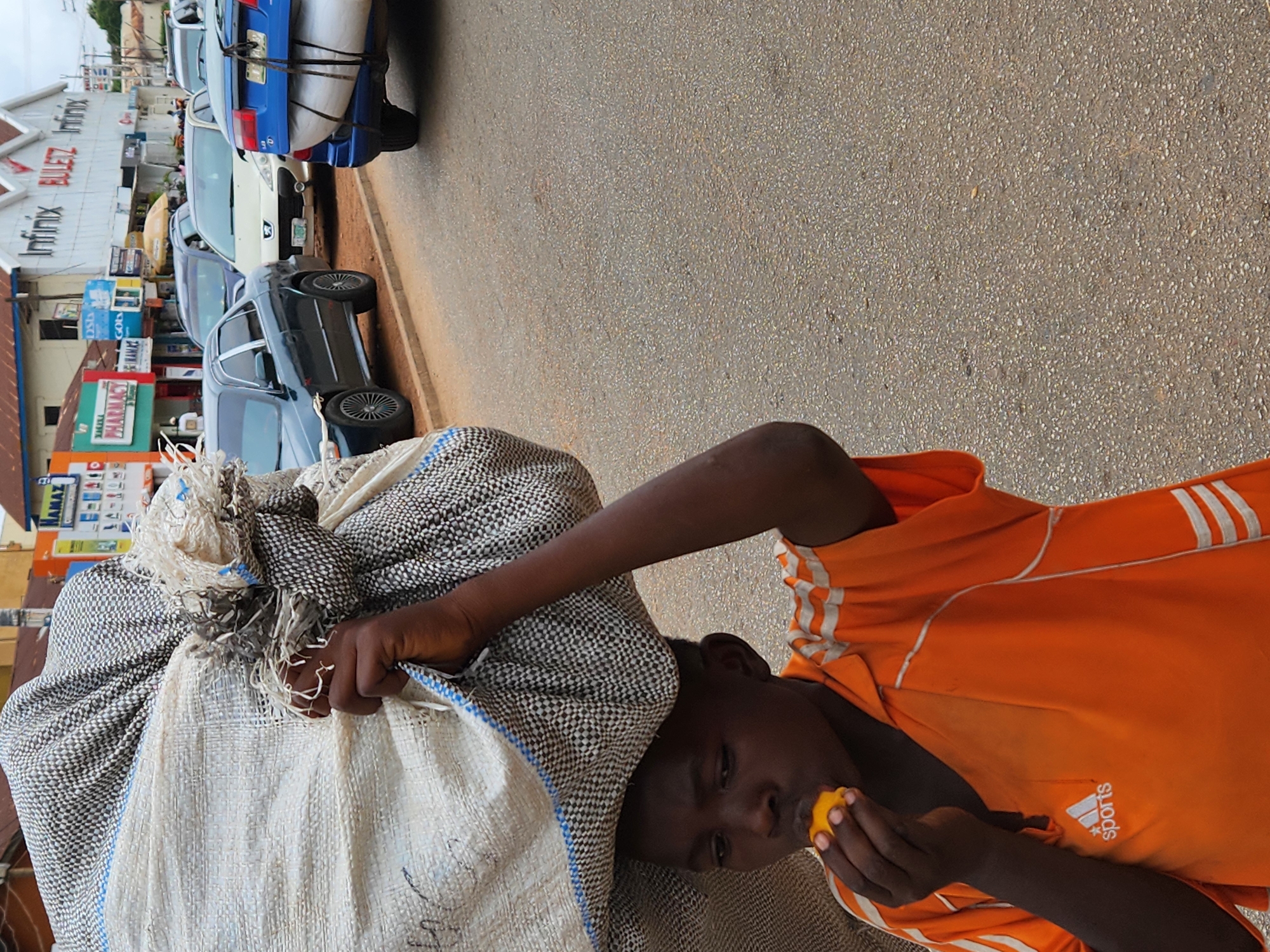 A young person, likely a male, wearing a bright orange t-shirt with white stripes and a "sports" logo, carries a large, light-colored, textured sack or bundle on their head. The person is eating a piece of yellow fruit, possibly a mango or orange, held in their right hand.
The scene is outdoors, on what appears to be a street or roadside in daylight. The road surface is asphalt. Along one side of the road, several cars are parked. These include a blue sedan with items strapped to its trunk, a white sedan, and a dark sedan with distinctive spoked wheels.
In the background, beyond the parked cars, several buildings are visible, suggesting a commercial area. Text visible on the buildings includes "Infinix" and "PHARMACY" on a green sign. Another sign has "RAMAZ" or similar text. The lighting suggests a bright, possibly sunny, day. The individual's expression is neutral, focused on eating the fruit. The image is rotated to a portrait orientation, making the street appear vertical.