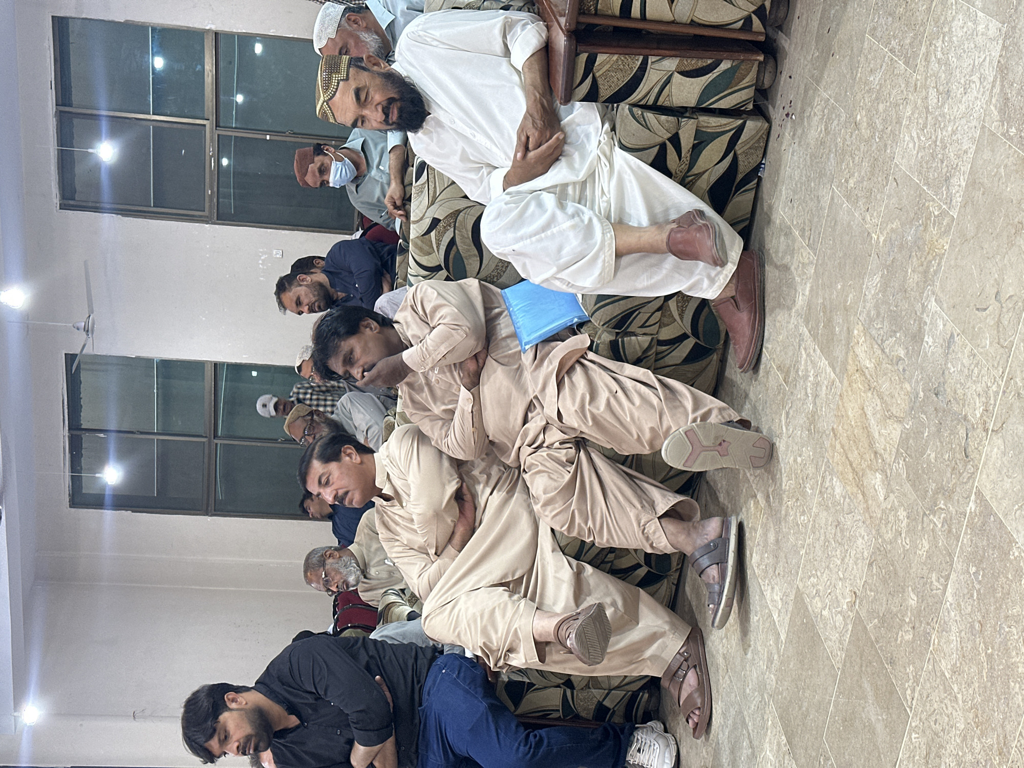 The image shows a group of men gathered indoors, possibly in Quetta City Tehsil, Pakistan, based on the clothing styles and features of the men. Some are sitting and appear to be resting or sleeping. The men are wearing a variety of traditional and casual clothing. The environment appears to be a simple indoor setting, potentially a gathering space in a home or community center, given the patterned sofa cushions and the presence of windows. There is no explicit action or interaction visible beyond the men's presence and posture.