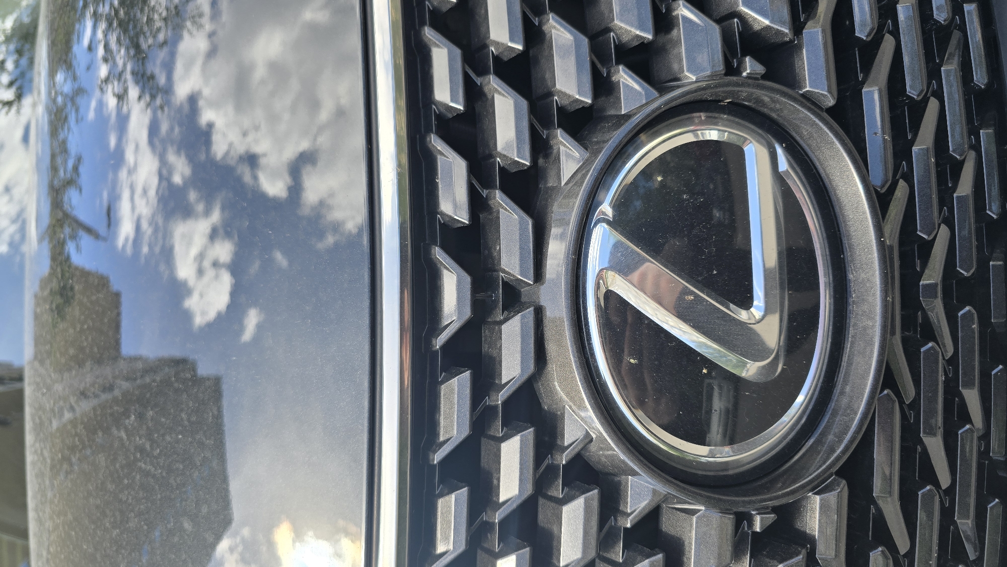 The image is a close-up, low-angle shot of the front grille and emblem of a dark-colored Lexus vehicle, presented with a vertical orientation.
The primary subject is the car's front, featuring its prominent, shiny metallic Lexus emblem, designed with a stylized "L" shape enclosed within a circular frame. The emblem reflects the bright sky and appears to have some dust or smudges. Surrounding the emblem is the vehicle's dark-colored grille, which has a complex, three-dimensional geometric pattern of interlocking, faceted shapes.
To the left of the grille, a significant portion of the car's highly reflective, glossy dark body is visible. This surface mirrors a bright blue sky with fluffy white clouds, indicating a clear, sunny daytime. Reflections of what appears to be tree branches or foliage are visible at the top, and an indistinct, light-colored building or architectural structure is reflected towards the lower-left. The reflective surfaces of both the car's body and the emblem show visible dust particles and smudges.
The scene conveys an outdoor setting on a sunny day, with urban or suburban elements suggested by the reflected building and trees. No human activity is depicted.