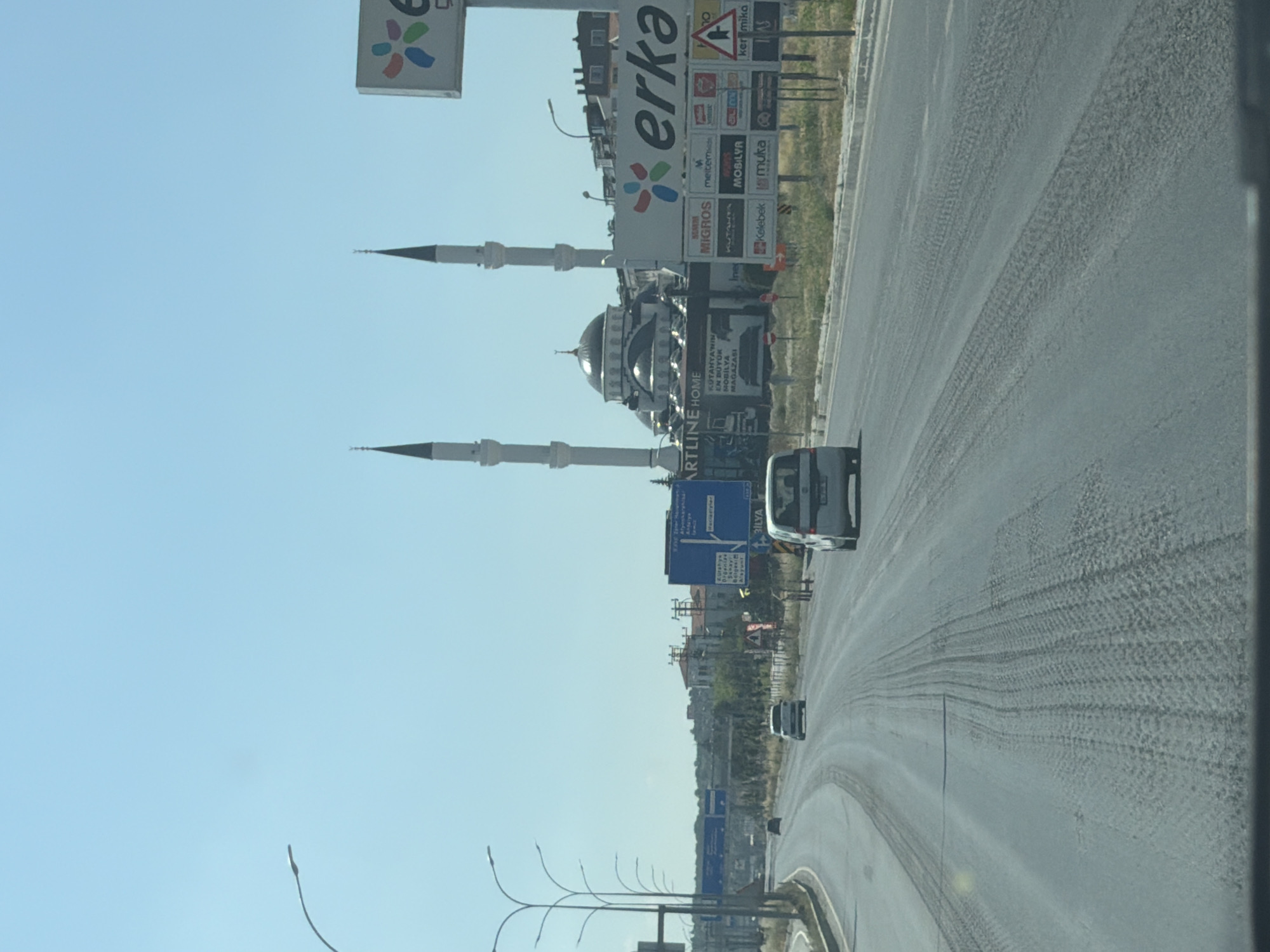 This is a medium-distance, eye-level, outdoor, daytime shot of a multi-lane road in Kütahya Merkez, Turkey. The road appears to be paved with asphalt and shows tire tracks from vehicles. A white car is visible in the right lane, and another white car is in the left lane. A mosque with two minarets is located in the background, to the left of a large billboard with advertisements. The sky is clear and blue. The scene appears to be a commercial or industrial area, given the presence of billboards and the architecture.