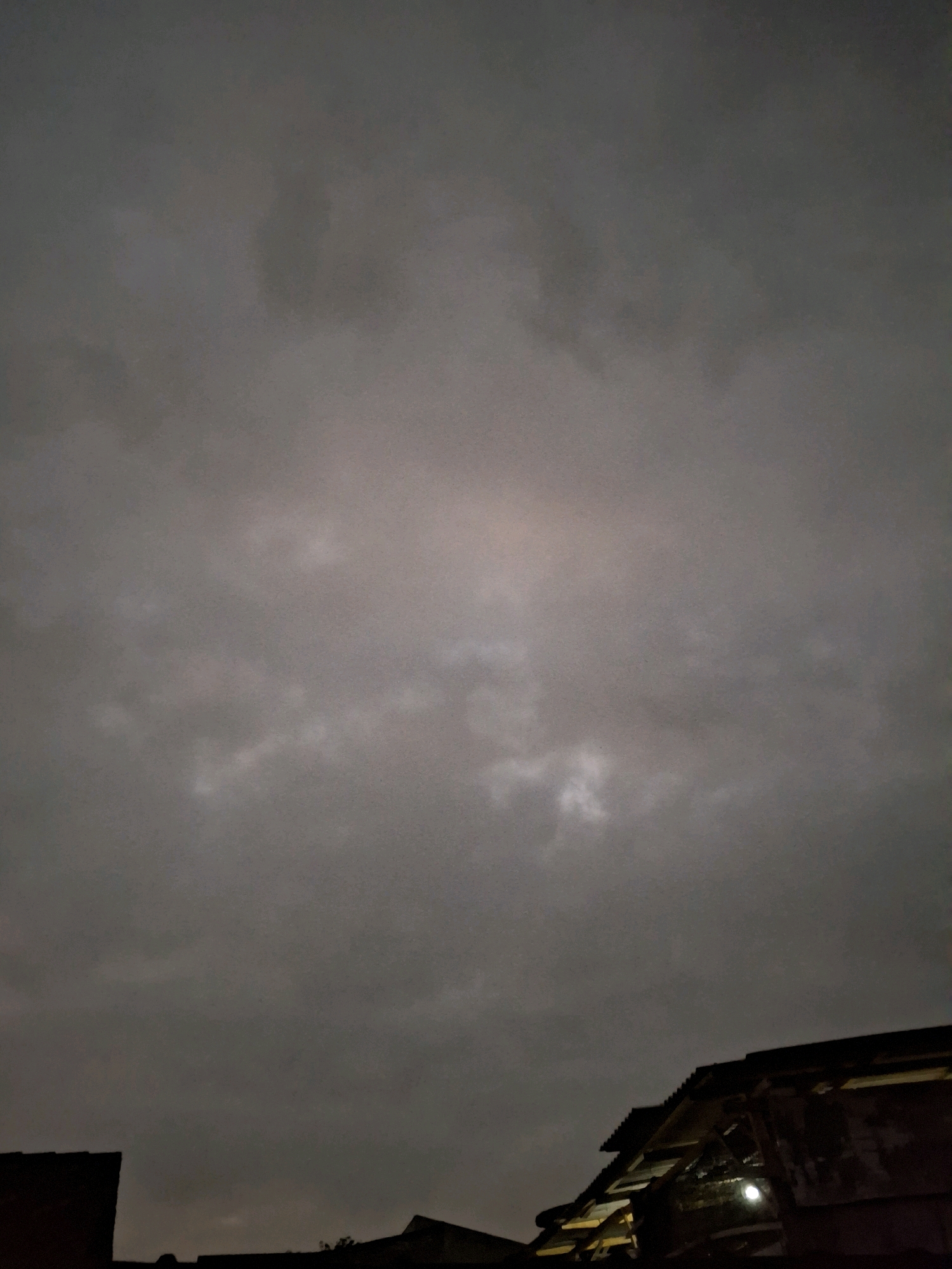 The image depicts a cloudy, overcast sky above the rooftops of buildings in Lumajang, Indonesia. The sky is predominantly gray with some lighter patches, suggesting a dim or twilight setting. The lower portion of the image shows the dark silhouettes of several building structures, including a tiled roof with visible wooden beams and a bright light source. The scene is devoid of any people or visible activity.