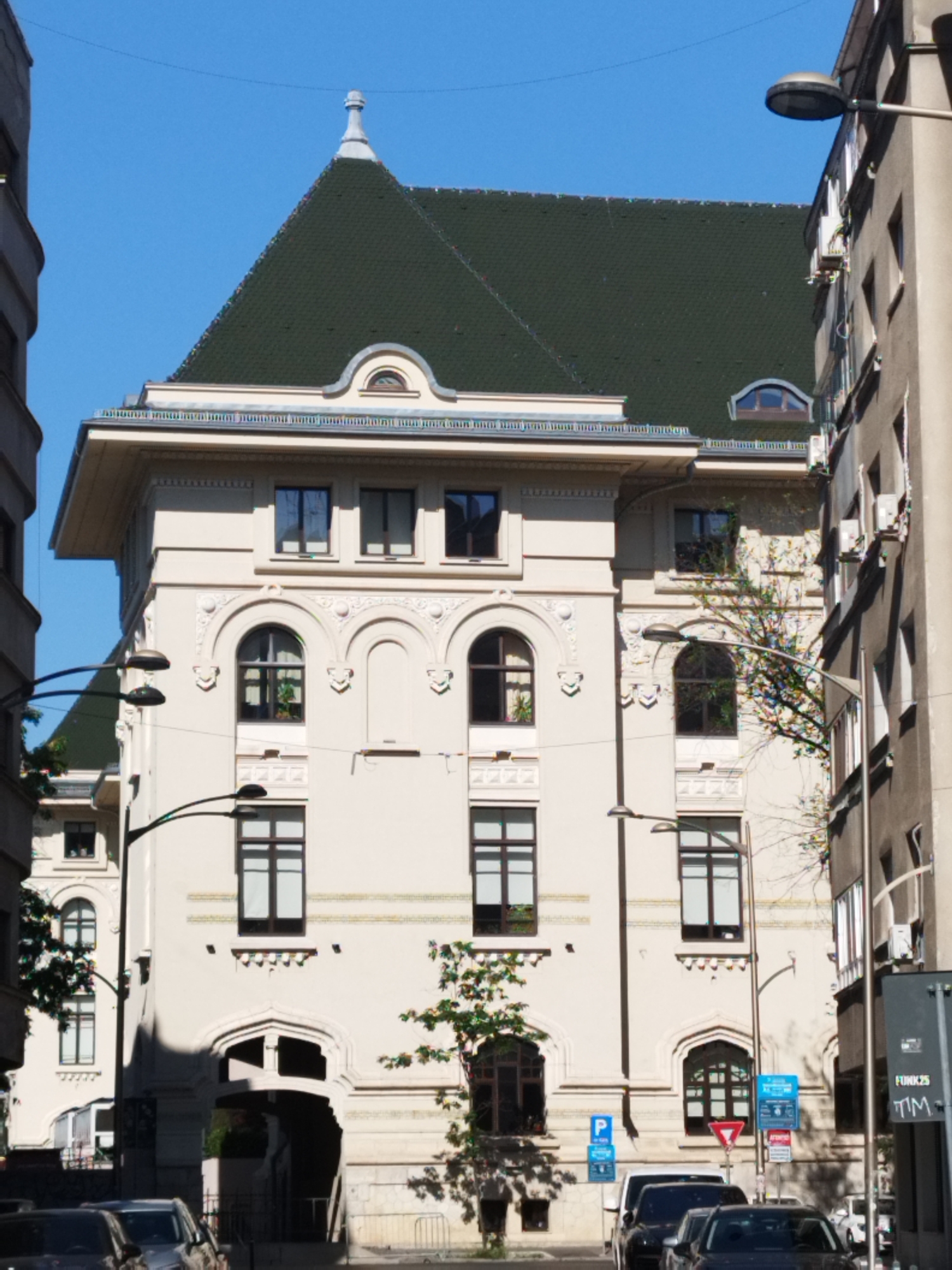 This is a street-level, eye-level medium shot of a beige building with an ornate facade and a dark green mansard roof, located in Bucharest, Romania. The building has several arched windows on its upper floors, some of which are tinted. A prominent arched entranceway is visible on the lower level.
Streetlights with modern, dark fixtures are positioned along the sidewalk in front of the building. Two cars are parked on the street, and a pedestrian street sign with "FUNK25 TIM" is mounted on a pole on the right side of the image. A small tree with sparse branches is visible growing in front of the building. The sky is clear and blue. The architecture suggests the building is likely historic, possibly a public building or an apartment complex from the early 20th century. The overall scene depicts a typical urban streetscape in Bucharest on a sunny day.