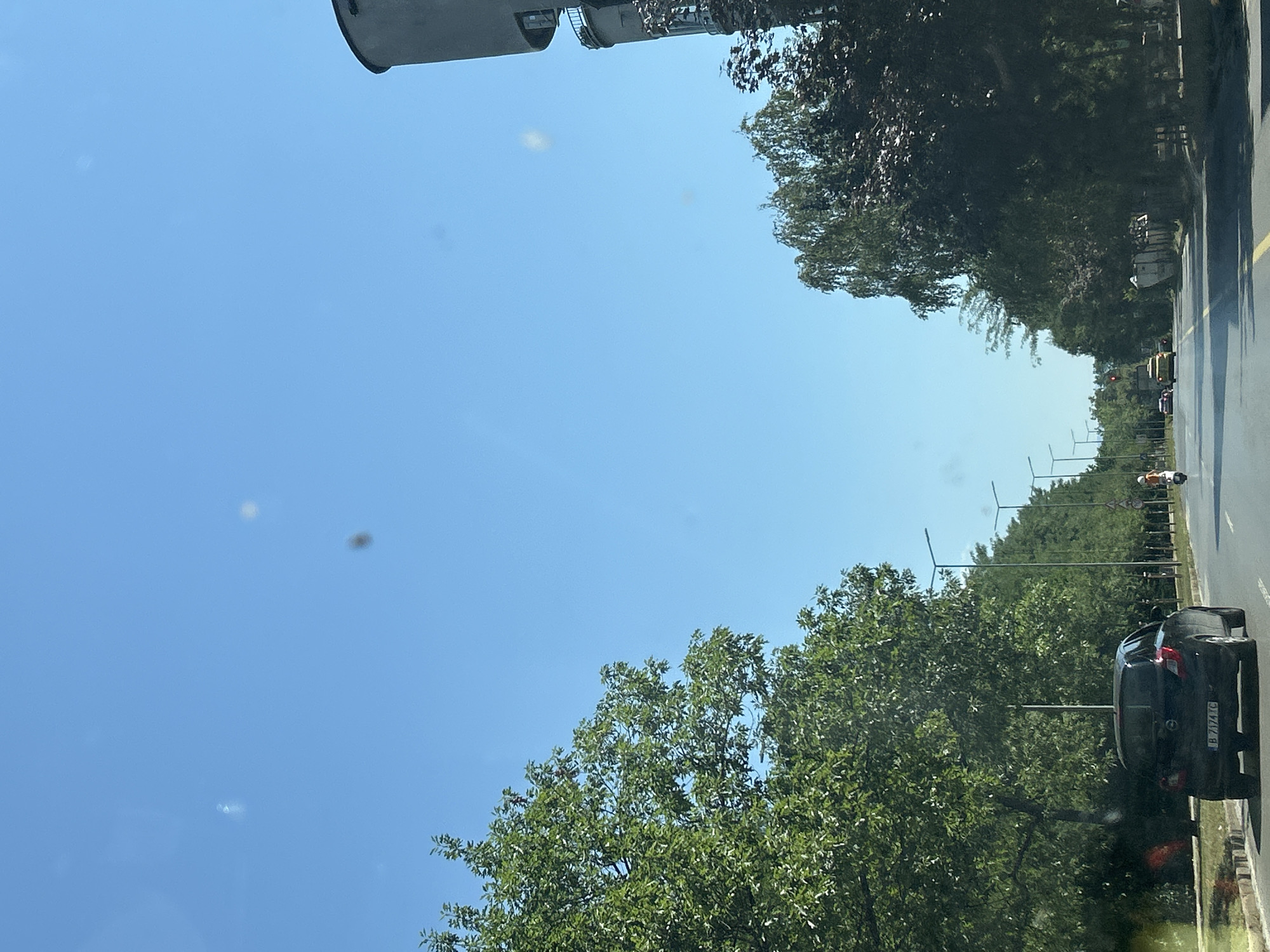 The image captures a view from a vehicle traveling down a tree-lined street in Sofia, Bulgaria, on a clear, sunny day. On the left side of the road, large green trees with dense foliage line the pavement. On the right side, the street also has trees, with a prominent grey cylindrical structure, possibly a water tower, visible in the upper right corner. The road itself is asphalt with yellow lane markings. Several vehicles are present, including a black Opel hatchback in the foreground with a Bulgarian license plate reading "B 7174 YC", a yellow car further down the road, and a scooter with a person on it. Streetlights are visible along the side of the road, and traffic signs, including a speed limit sign of 20, are also present. The sky is a bright, clear blue with a few faint, blurry spots, possibly dust or lens artifacts.