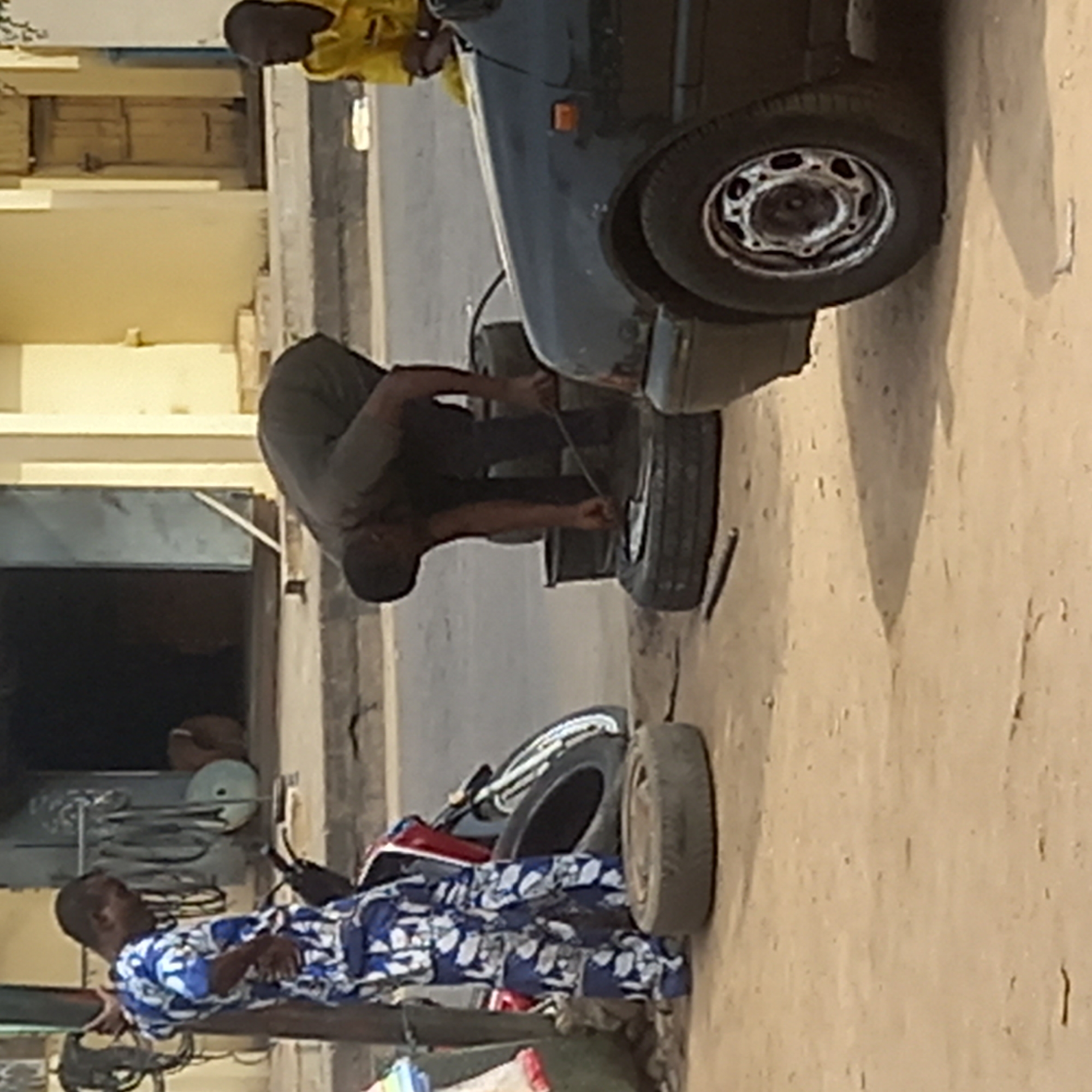 The image depicts an outdoor scene in Sardauna, Nigeria, showing tire repair work. A man in a grey shirt is kneeling and using a tool to work on a tire attached to a dark-colored car. Two spare tires are on the ground nearby. Another man in a patterned blue and white shirt is reclining in the lower left corner of the image. A man in a yellow shirt is partially visible in the upper part of the frame. The environment appears to be an urban roadside or workshop area.