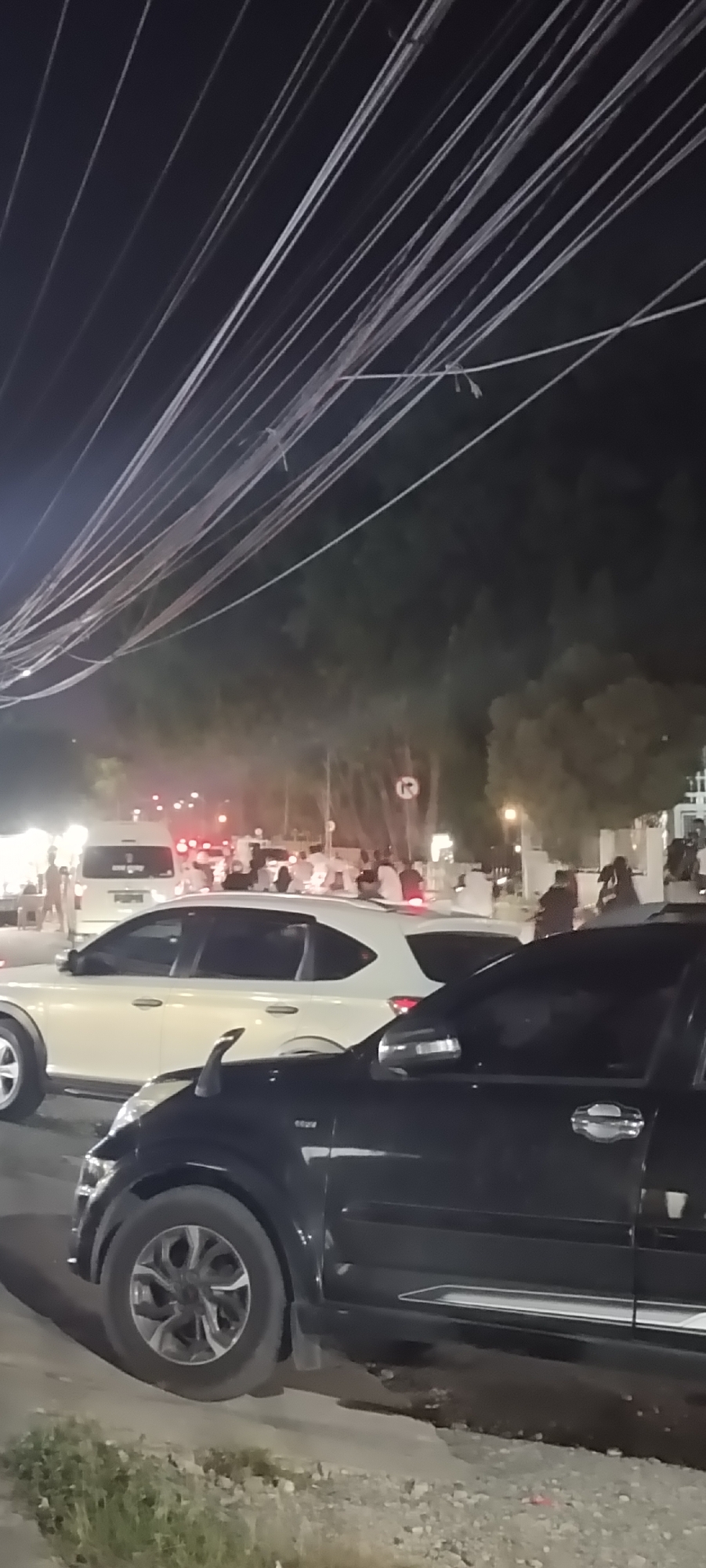 The image shows a street scene at night in Lhokseumawe, Indonesia. A black car is parked on the right side of the frame, partially blocking the view. Behind it, there is a white car parked in front of a crowd of people gathered near a lit area. Overhead, a network of power lines crisscrosses the sky. The background suggests a public space with trees and a building in the distance.