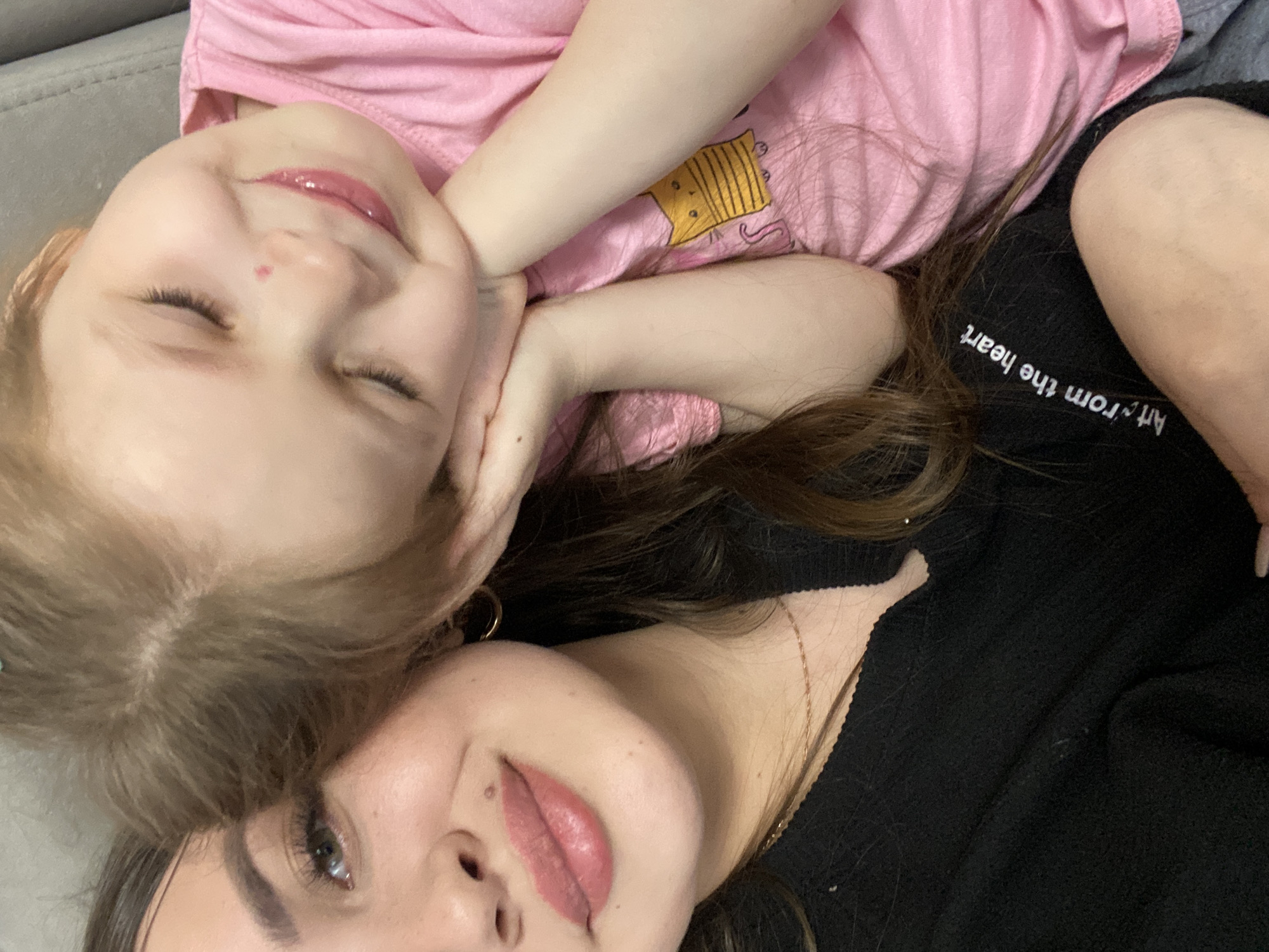 Two individuals, likely a mother and daughter, are in close proximity, possibly lying down. The daughter, a young child with light hair, is wearing a pink shirt with a cartoon graphic. The mother has dark hair and is wearing a black top with the words "Art from the heart" printed on it. The background appears to be a gray upholstered surface, possibly a couch or bed. There is no indication of the location being in Chaikovsky, Russia, in the image.