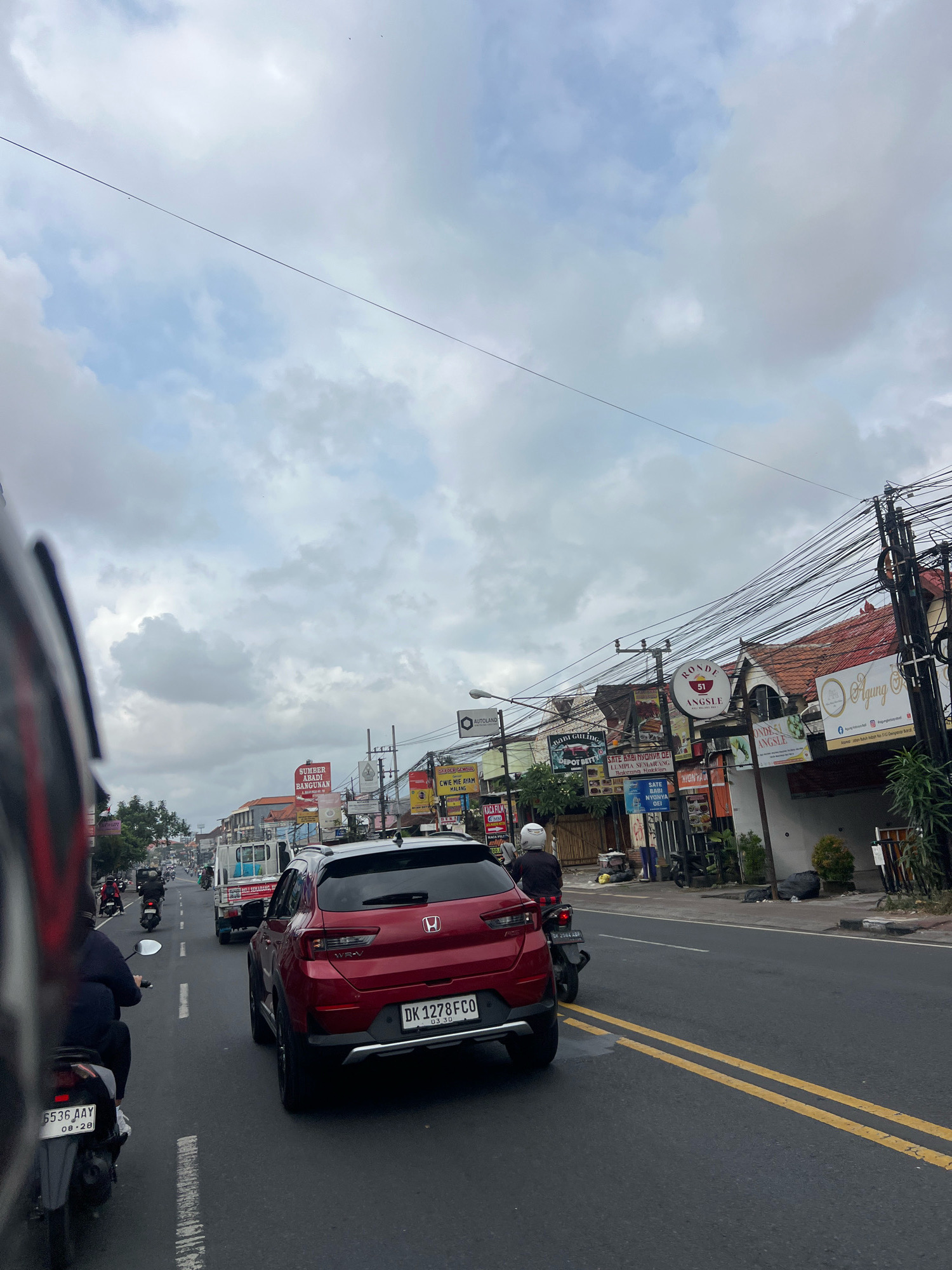 The image depicts a street scene in Denpasar, Indonesia, characterized by moderate traffic and commercial establishments. The perspective is from behind a motorcycle, looking forward along a two-lane road. A red Honda WR-V SUV is in the foreground, in the right lane, with its license plate visible as DK 1278 FCO. To the left of the SUV, a motorcycle with license plate 5536 AAY is also visible. Several other motorcycles and a white truck are further ahead in traffic. The sidewalks are lined with businesses, indicated by various signs advertising food, services, and shops, some with Balinese script. Buildings have tiled roofs, and a dense network of electrical wires is strung overhead. The sky is overcast with clouds.