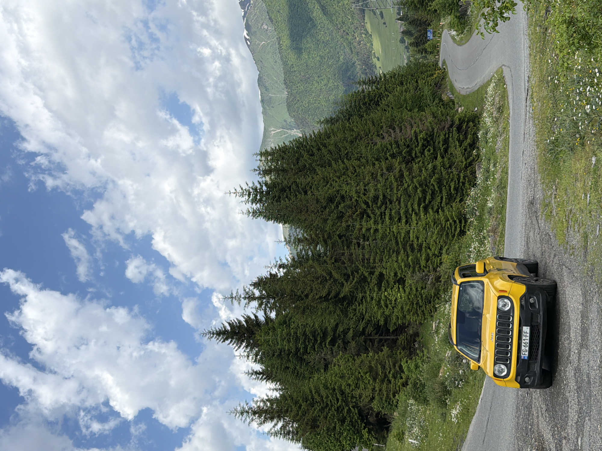 A yellow Jeep Renegade is parked on a winding mountain road in Mestia, Georgia. The Jeep is facing the camera, with its headlights on. The road is asphalt and curves to the right, leading up a hill. To the left of the road is a dense forest of tall, dark green pine trees. To the right, the road continues to ascend, with the landscape opening up to reveal rolling green hills and snow-capped mountains in the distance. The sky is a bright blue with fluffy white clouds. The image appears to be taken during the daytime, likely in the late spring or summer, given the lush greenery and visible snow on the peaks. There are no people visible in the image. The overall scene depicts a scenic mountain landscape with a vehicle on a road.