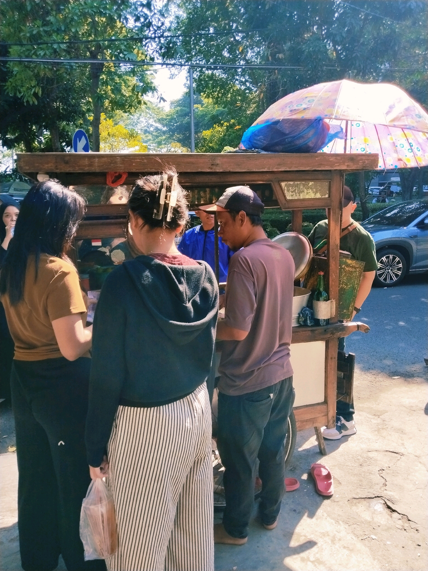 In a sunny street scene in Surabaya, Indonesia, a food cart is the central focus. Three individuals are gathered around the cart, appearing to be engaged in a transaction or conversation. One person, wearing a purple t-shirt and dark pants, stands with their back to the camera, facing the cart. To their left, a person in a dark hoodie and vertically striped pants holds a plastic bag containing what appears to be food. Another person, in a brown t-shirt and dark pants, stands on the far left, also facing the cart. A fourth individual, wearing a blue shirt, is partially visible behind the cart. The cart itself is wooden with an umbrella providing shade, displaying various items within. A blue car is parked on the right side of the frame, and lush green trees line the background. The ground is paved with concrete.