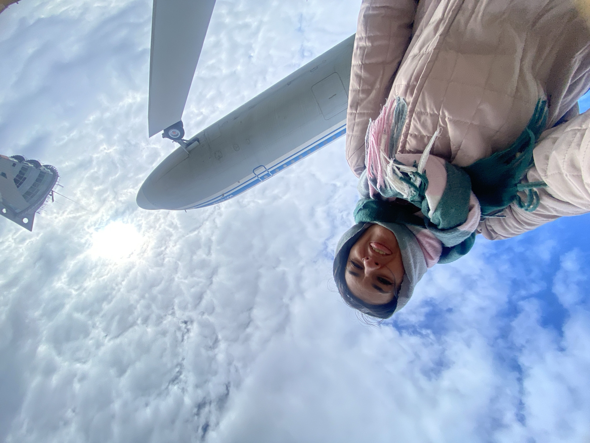 The image presents a striking low-angle selfie, capturing a person and a large aircraft against a dynamic sky. In the foreground, at the bottom right, a person with dark hair, wearing a light brown quilted hooded jacket and a colorful scarf (pink, green, and white), is visible. The individual is looking towards the camera with a gentle smile or smirk, showing some teeth.
Dominating the upper and central parts of the frame is the underside of a substantial grey aircraft fuselage, accented with blue stripes along its side. A portion of a propeller or wing and a landing gear wheel assembly are visible in the upper right corner, indicating a turboprop or jet aircraft. Visible Cyrillic text on the fuselage reads "ИЛ-62М" (IL-62M), identifying it as an Ilyushin Il-62M, and a partial registration "RA-865..." is also discernible. On the far left, a blurry, angular structure, possibly part of a building or another large object, is also present.
The setting is outdoors, under a bright, partly cloudy sky. The sun is prominent in the upper left quadrant, radiating light through the clouds and creating a bright glare. Patches of clear blue sky are interspersed among the fluffy clouds. The wide-angle lens used for the photograph creates a distinctive, somewhat distorted perspective, emphasizing the scale of the aircraft and the expansive sky. The overall impression is of a cheerful moment captured at what appears to be an aviation display or museum, with the person enjoying a close view of the historic aircraft.