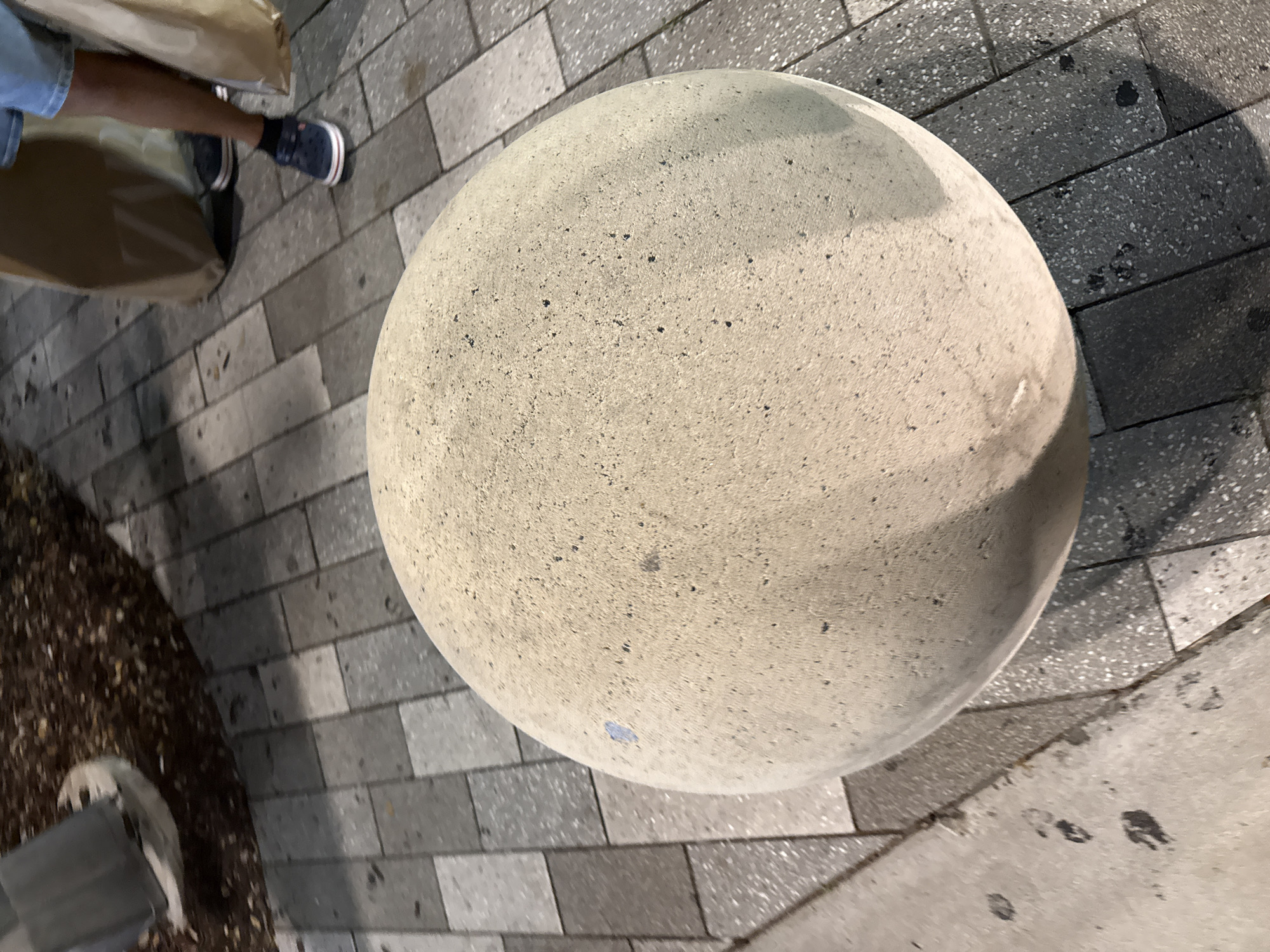 The image provides an overhead view of an outdoor urban setting, likely a paved area in Aventura, USA. A large, light-colored concrete sphere, possibly a decorative bollard, is prominent in the center-right, featuring a rough, speckled texture with dark aggregate. A small, faint blue mark is visible on its lower side.
To the left, the lower legs and feet of a person are visible, wearing light blue shorts and dark blue shoes with white soles. Two brown paper shopping bags rest near their legs, suggesting recent shopping activity. The person appears to be standing or walking on the paved surface.
The ground is covered with rectangular and square tiles in varying shades of grey, some exhibiting a fine speckled pattern. Shadows, notably from the concrete sphere, are cast across the pavement, indicating the presence of a light source, suggesting late afternoon or early evening lighting conditions. In the bottom-left corner, a patch of dark, loose material, possibly mulch or soil, with scattered light debris, is visible alongside a low, dark grey structure. No discernible text is present in the frame.