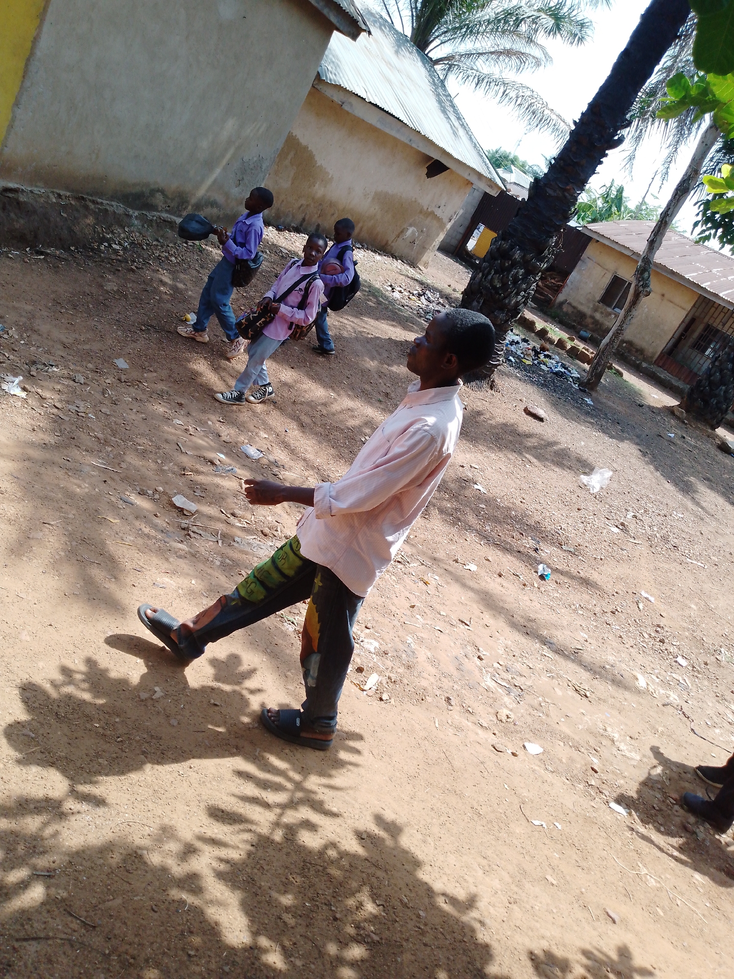 The image shows a dusty outdoor scene with several people and buildings. In the foreground, a man with dark hair and a light pink shirt stands at an angle to the camera. He wears dark pants with some kind of design and sandals. Behind him and slightly to the left, three younger people, possibly students, walk in the same direction. The first one carries a round object, possibly a ball or a hat. The second has a shoulder bag, and the third carries a backpack. All three appear to be wearing similar school uniforms. In the background, there are simple buildings with corrugated metal roofs and unfinished or earthen walls. A tall palm tree is prominent to the right, casting shadows on the ground. Litter and debris are scattered across the dirt ground, suggesting a somewhat rural or underdeveloped area.
