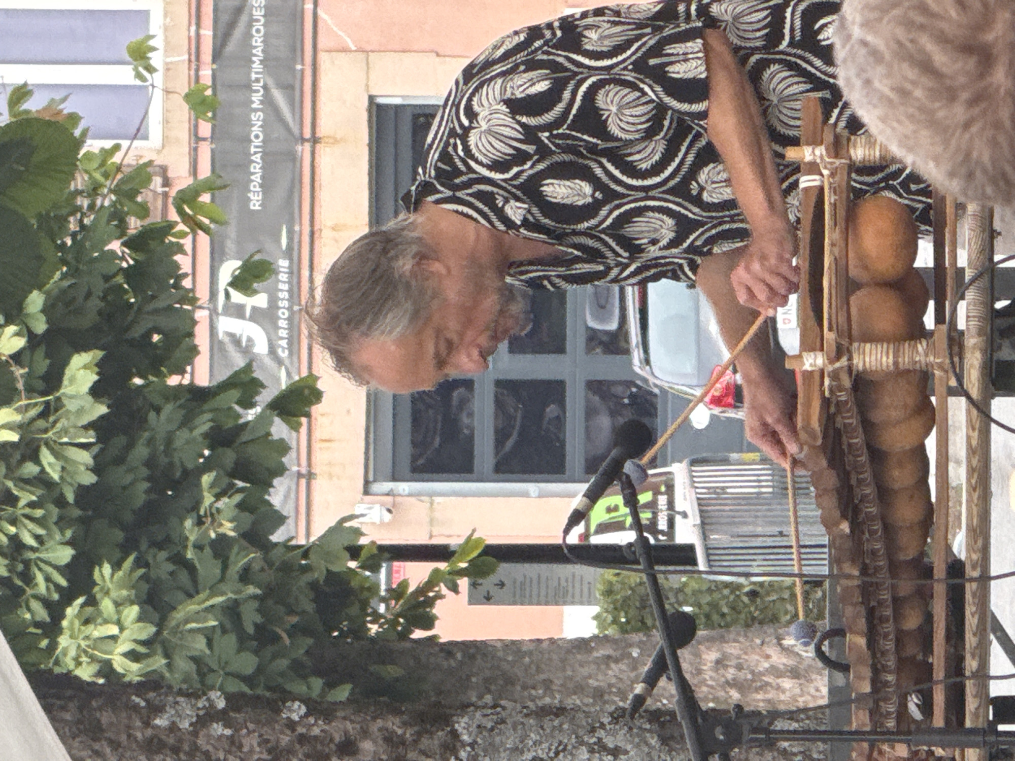 In the city of La Chaux-de-Fonds, Switzerland, a middle-aged man with graying hair and a goatee is playing a balafon, a type of wooden xylophone. He is wearing a black short-sleeved shirt with a white tropical leaf pattern. His hands are positioned over the instrument, holding two mallets, one of which is striking a wooden bar. The balafon has a wooden frame with gourds attached underneath to act as resonators. Microphones are set up on stands in front of the instrument to amplify the sound. In the background, there is a tree with green leaves and a building with a sign that reads "JR CARROSSERIE RÉPARATIONS MULTMARQUES." A white vehicle is partially visible behind the balafon. The scene appears to be an outdoor performance.