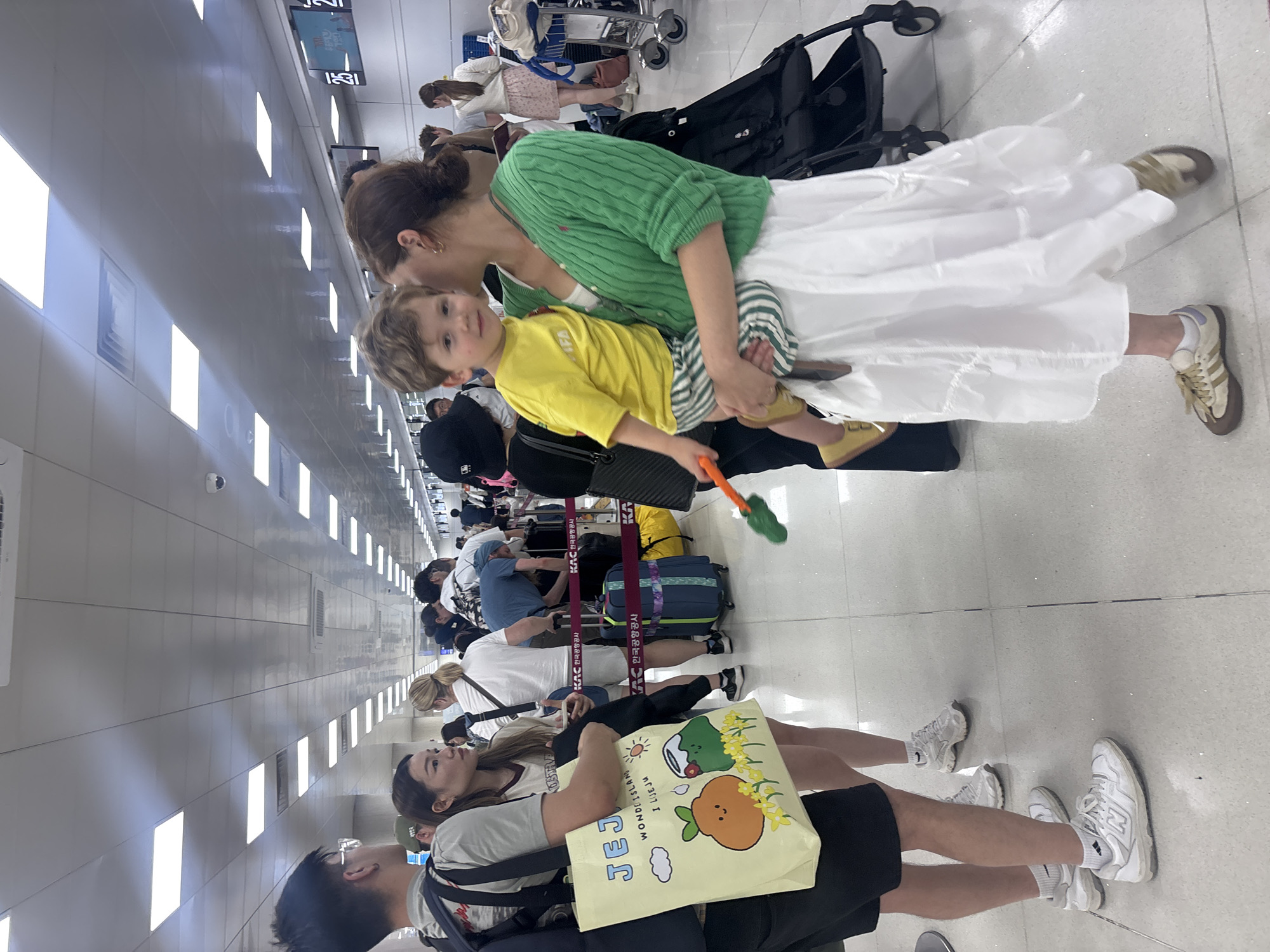 The photograph captures a busy airport scene, likely in Jeju, South Korea, given the visible "JEJU" and "한국공항공사" (Korea Airports Corporation) text on bags and barriers. A woman is holding a young child, who is looking towards the camera with a curious expression. The woman is wearing a green knitted cardigan over a white dress, and the child is dressed in a bright yellow t-shirt and striped shorts.
In the background, a line of people are standing, appearing to be waiting. Several pieces of luggage, including suitcases and trolleys, are visible, suggesting travel. Some individuals are holding or interacting with their belongings. The environment appears to be indoors, with bright, artificial lighting illuminating the tiled floor and the long, utilitarian walls. The overall atmosphere seems to be one of travel and transit, with people engaged in the common activities of an airport. The image is taken from a slightly elevated angle, looking down at the subjects and the surrounding environment.