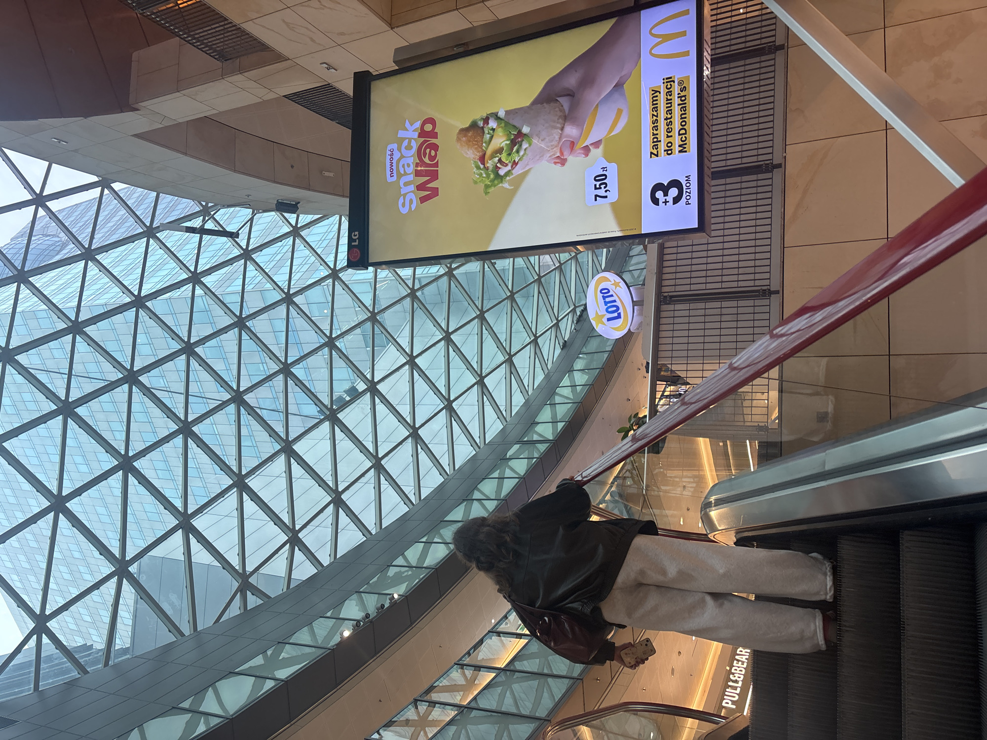 A slightly upward-angled shot captures a scene inside a modern shopping mall, likely in Warsaw, Poland. The focus is on a person standing on an escalator going down. She is wearing light-colored trousers, a dark jacket, and is carrying a brown bag.
Above the escalator, an advertisement for McDonald's "Snack Wrap" is clearly visible with Polish text, including the price of 7.50 zł, and the text "Zapraszamy do restauracji McDonald's." A Lotto sign is nearby.
The background showcases an architectural design with a large triangular glass structure. A "Pull&Bear" store sign is visible near the escalator's end.