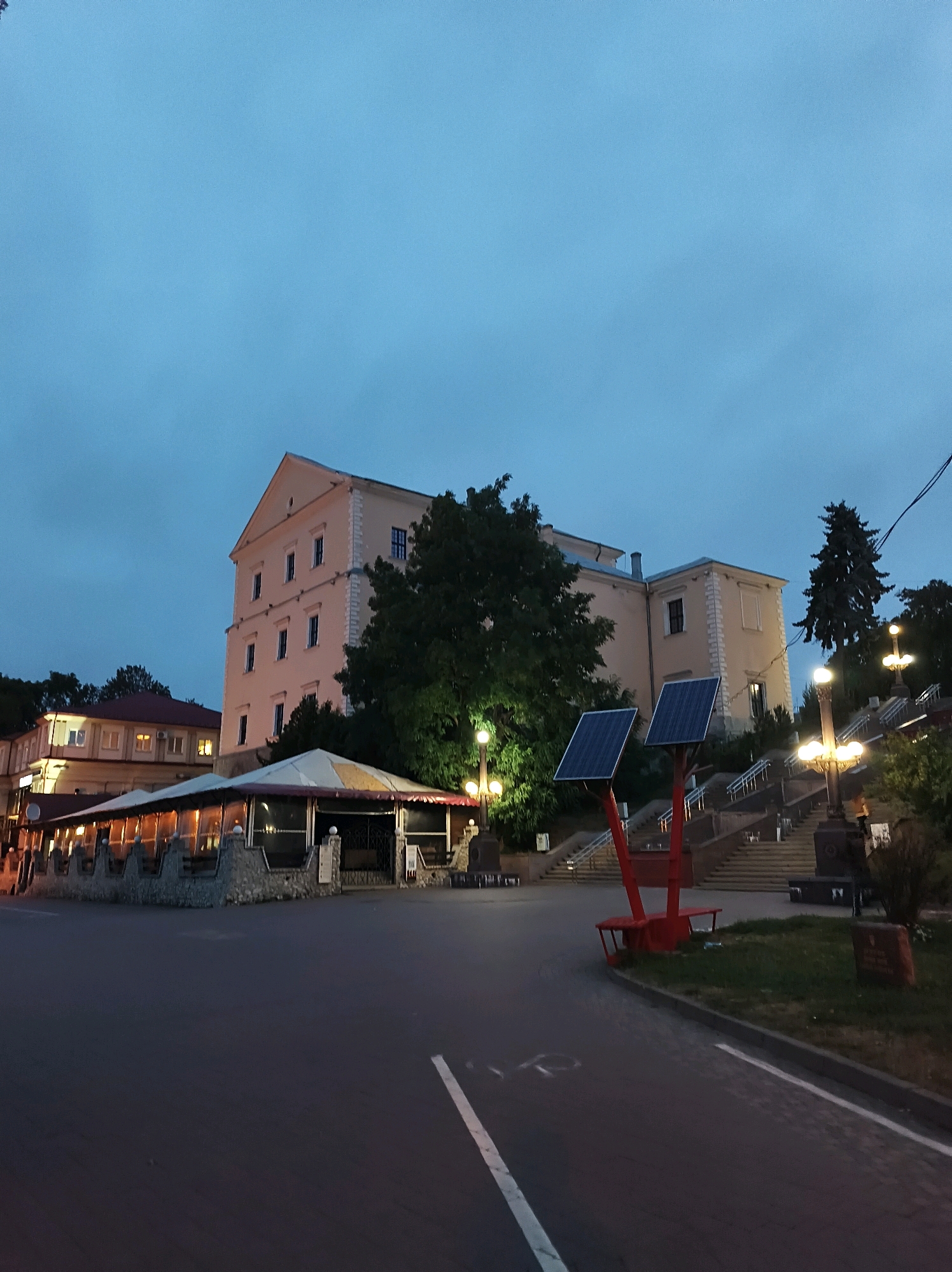The image shows a building on a hill with a staircase leading up to it. There are solar panels on the left side of the staircase and streetlights along the path. In the foreground, there is a road and a restaurant with a covered outdoor seating area. The sky is overcast, and it appears to be evening. The building on the hill is likely a historical landmark or administrative building. The restaurant is a modern establishment. The overall scene suggests a public space in the city of Ternopil, Ukraine, where historical architecture meets contemporary infrastructure. There are no people visible in the image.