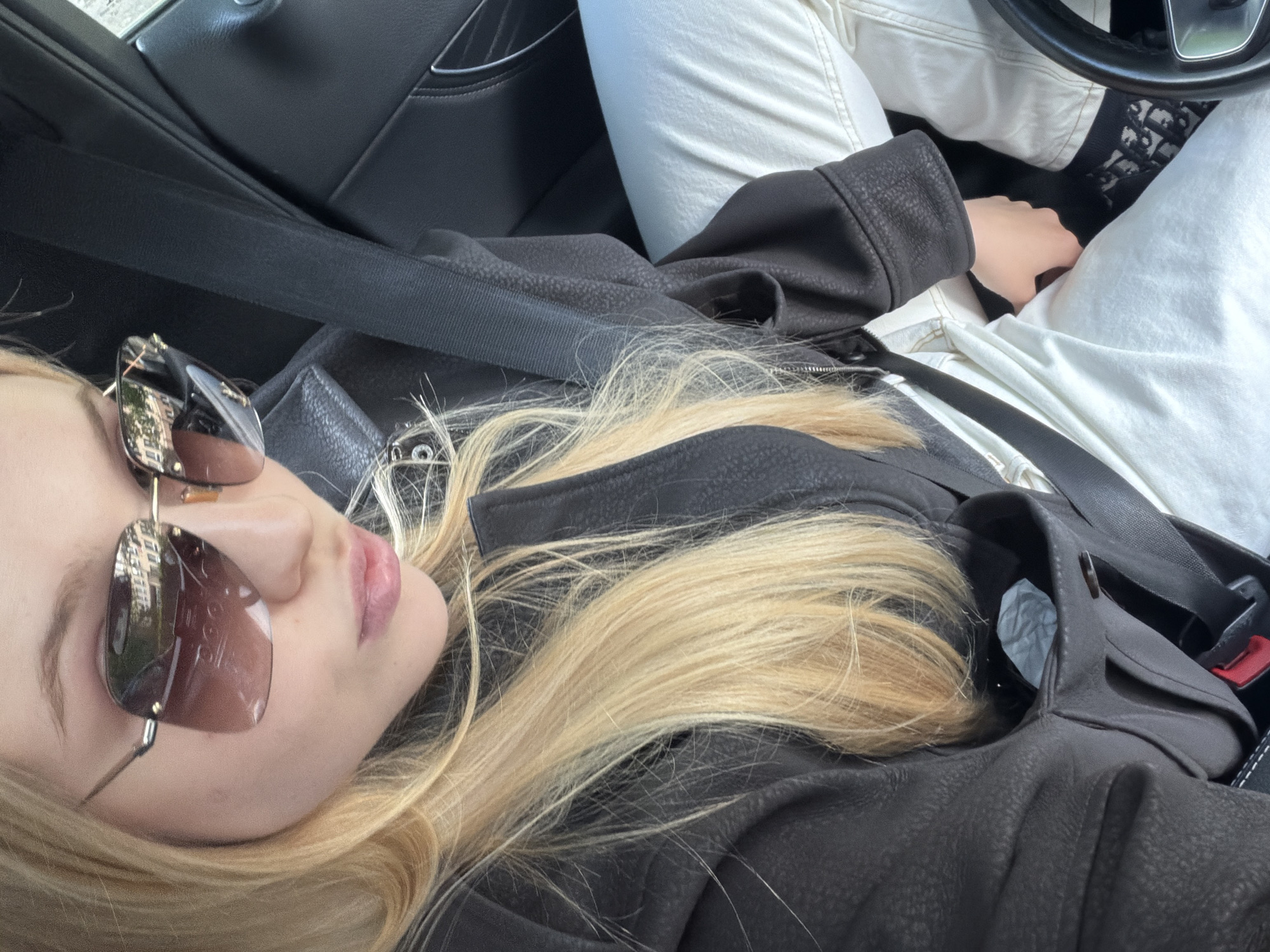 A woman with blonde hair and sunglasses sits in a car, wearing a black leather jacket and a seatbelt. She is wearing white pants and has a black and white patterned item near the steering wheel. The location is Unknown, Unknown.