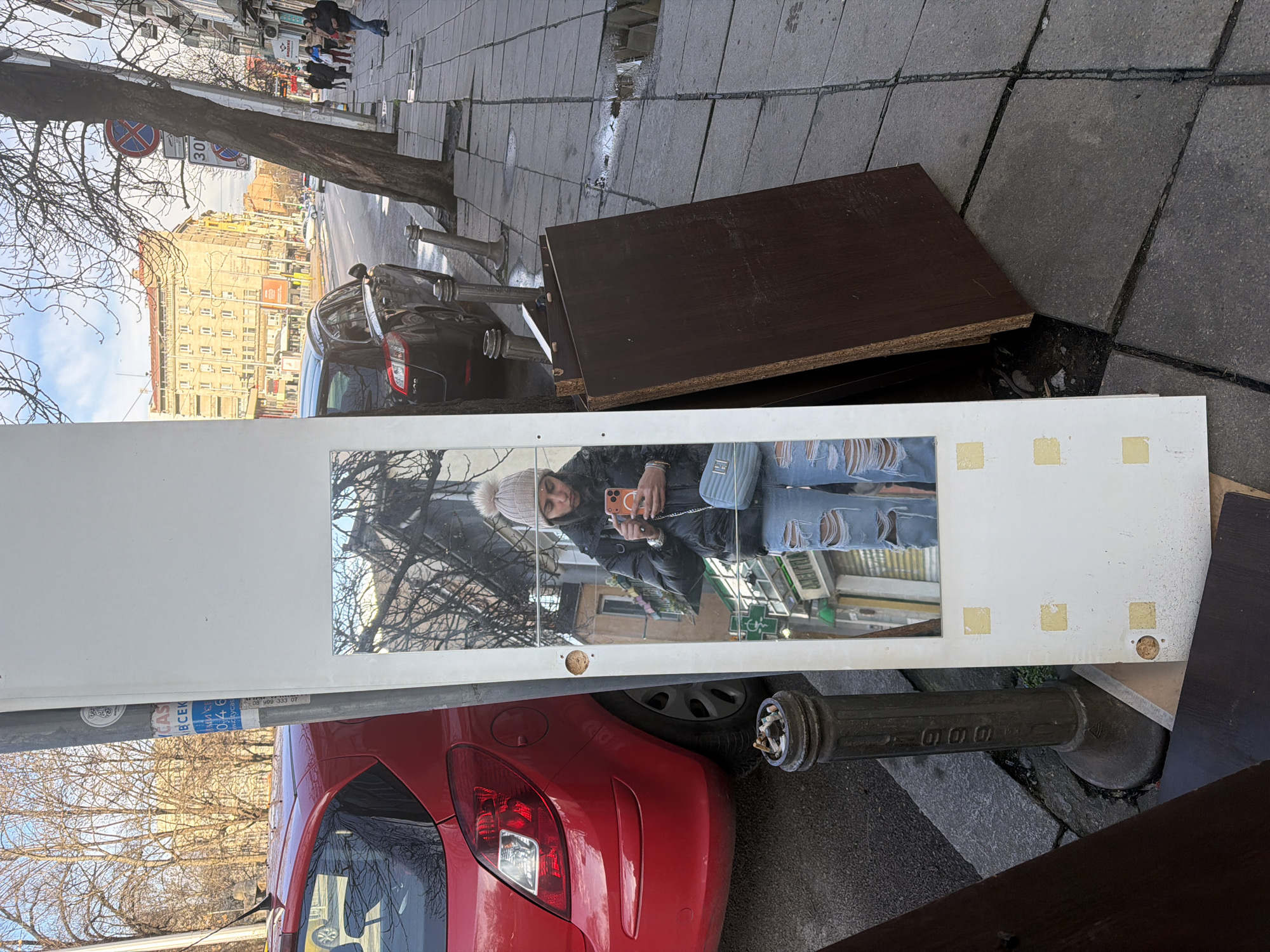 An urban street scene in Sofia, Bulgaria, features a white, vertically oriented furniture panel with an inset mirror leaning on a grey paved sidewalk. The mirror reflects a person, likely female, wearing a dark puffy jacket, ripped blue jeans, and a light-colored knit beanie, holding an orange-cased smartphone to take a selfie. A light blue quilted purse is visible across her body.
To the left of the white panel, the rear portion of a red hatchback is parked on the asphalt street, next to a metal bollard repurposed as an ashtray filled with cigarette butts. Behind the car, a light grey utility pole bears a sticker with "СОФИЯ ВСЕКИ" (Sofia Every) and "МЕДИА" (Media) in Cyrillic, confirming the location. Bare tree branches are visible overhead.
To the right of the white panel, several dark brown particleboard planks are stacked on the sidewalk. A black sedan is parked on the street beyond these planks. A large, bare-branched tree stands on the sidewalk, with two street signs attached: a red-bordered circular "No Stopping" sign and a white-bordered circular "30 km/h speed limit" sign. Metal parking bollards delineate the curb.
In the background, a multi-story building with a yellowish facade dominates the street view, characteristic of Bulgarian urban architecture. Other cars are visible on the street, and a few pedestrians walk on the far sidewalk. The sky is clear blue with some clouds, and the bare trees suggest a winter or early spring setting.