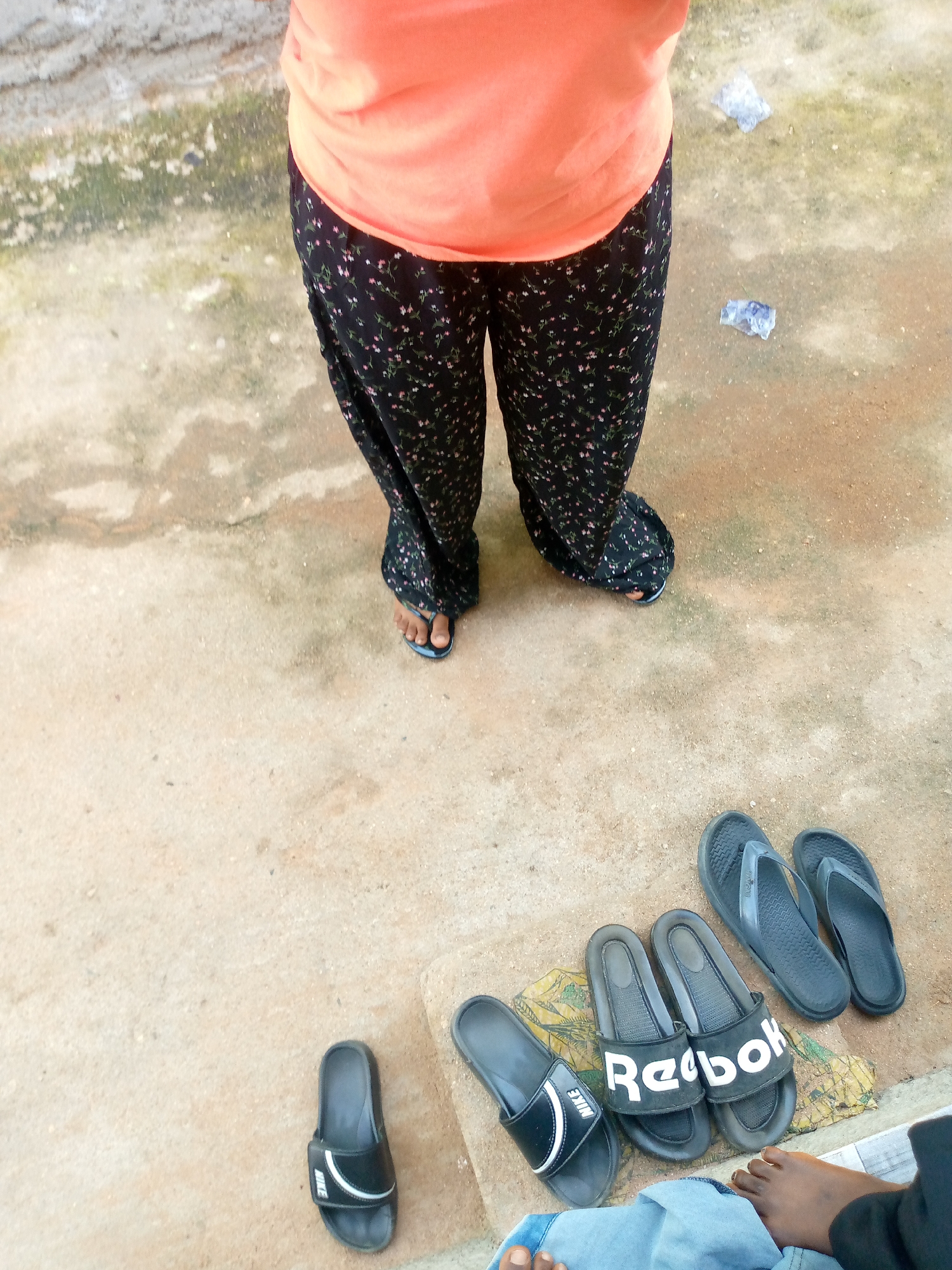 In Jauro Yinu, Nigeria, a person wearing an orange top and black floral patterned pants stands on a concrete surface. Their feet are adorned with blue flip-flops. In front of them, on the ground, are several pairs of black sandals. One pair is labeled "Nike," and another is labeled "Reebok." The ground is a mix of concrete and dirt, with some green moss and patches of white and blue plastic scattered around.