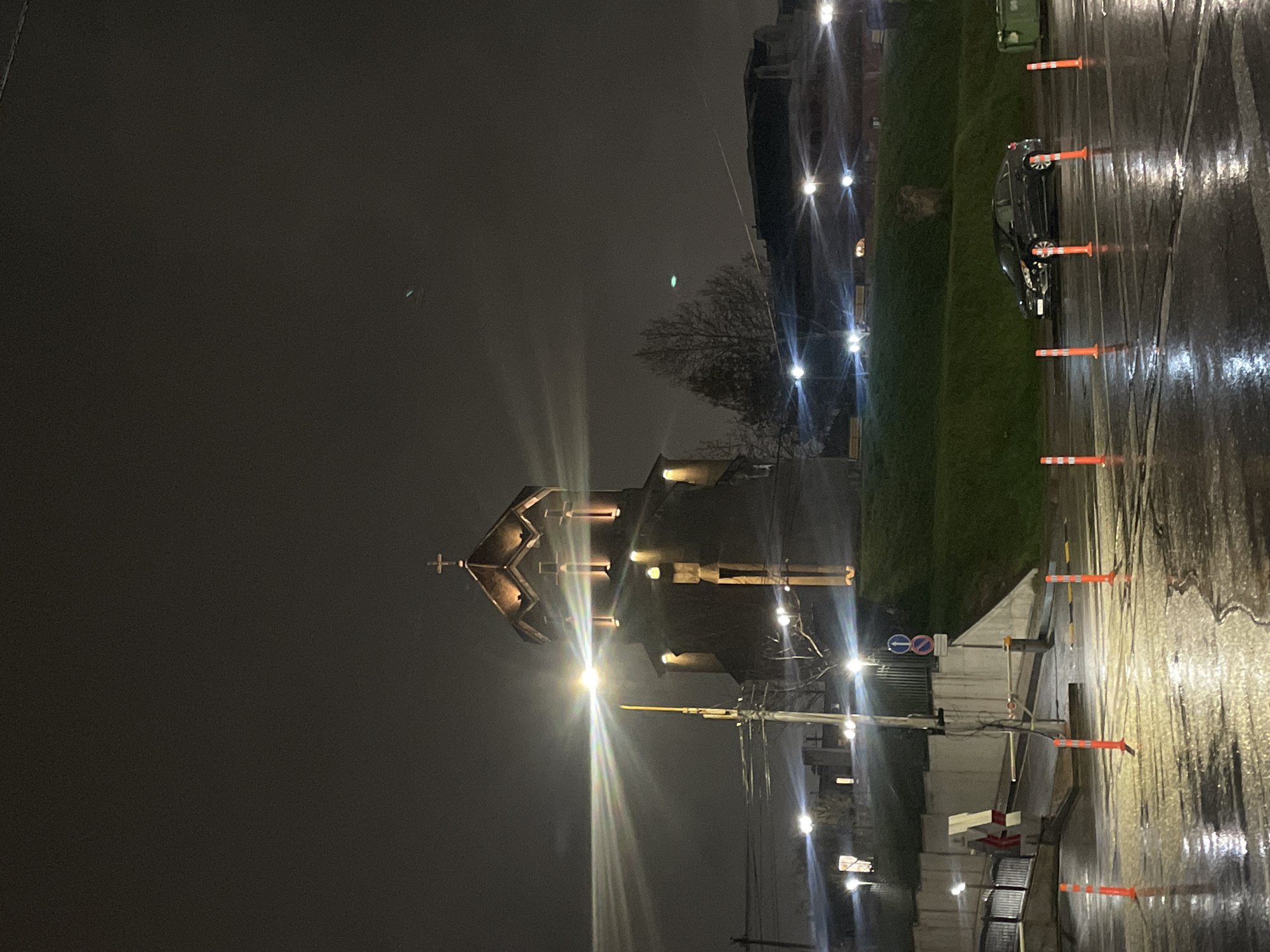 The image depicts a nighttime scene in Yerevan, Armenia. A church with illuminated windows and a cross on its roof is the focal point. Streetlights cast a bright glow. Wet pavement reflects light. Orange traffic cones line the side of the street. A dark-colored car is parked. Trees are visible. The sky is overcast. Utility poles and wires are present. A building is on the right, with lit windows. A road sign can be seen.