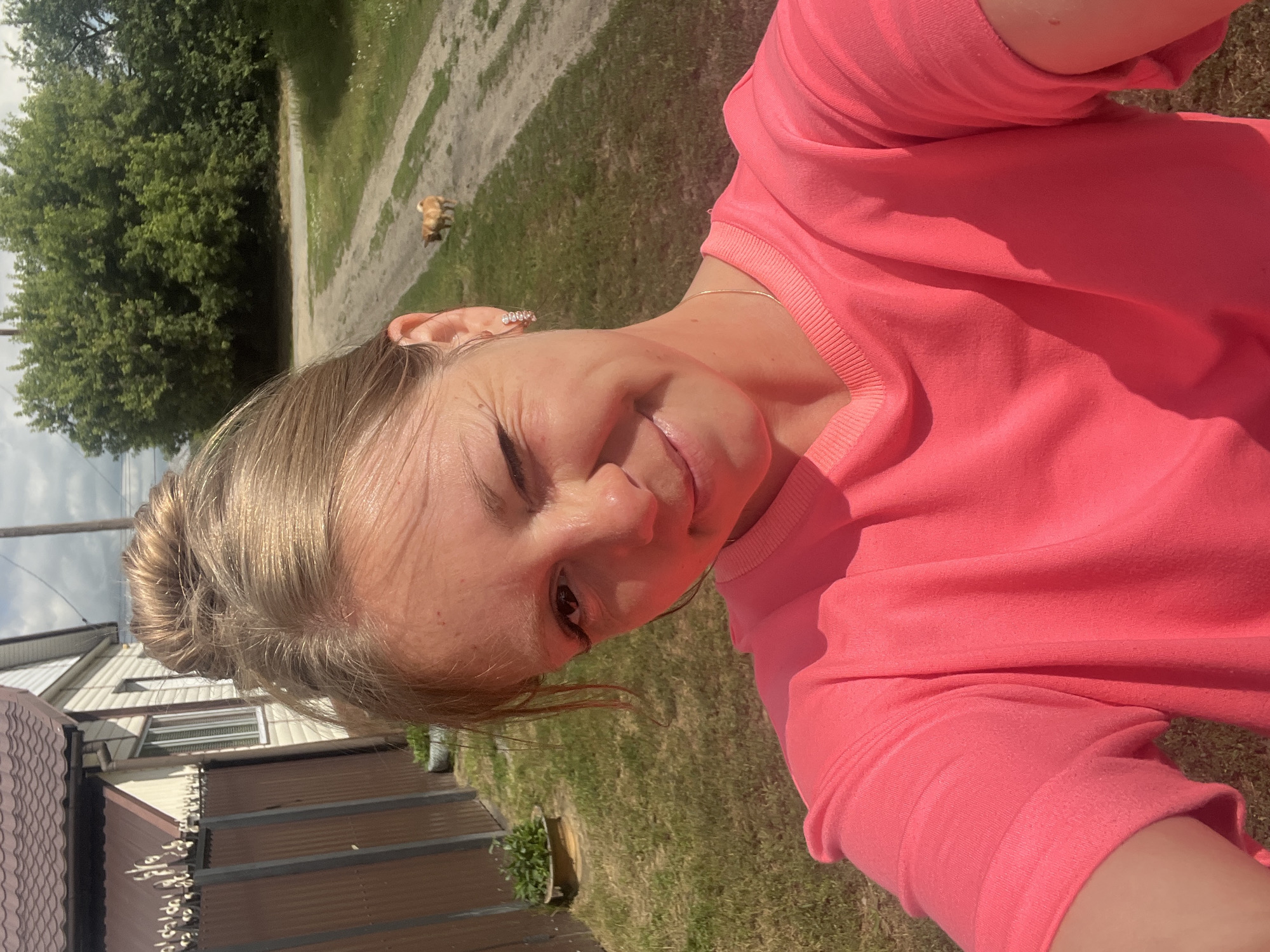 A young woman with blonde hair pulled into a bun winks at the camera while taking a selfie outdoors. She is wearing a bright pink long-sleeved shirt. In the background, a dirt road leads past a grassy area and a line of trees. A small, light-colored dog is visible walking on the road in the distance. To the left of the image, part of a brown fence and a house with a brown roof are visible. The overall setting appears to be a rural or suburban area in Сухринское, Russia, given the presence of a house, fence, and unpaved road.
