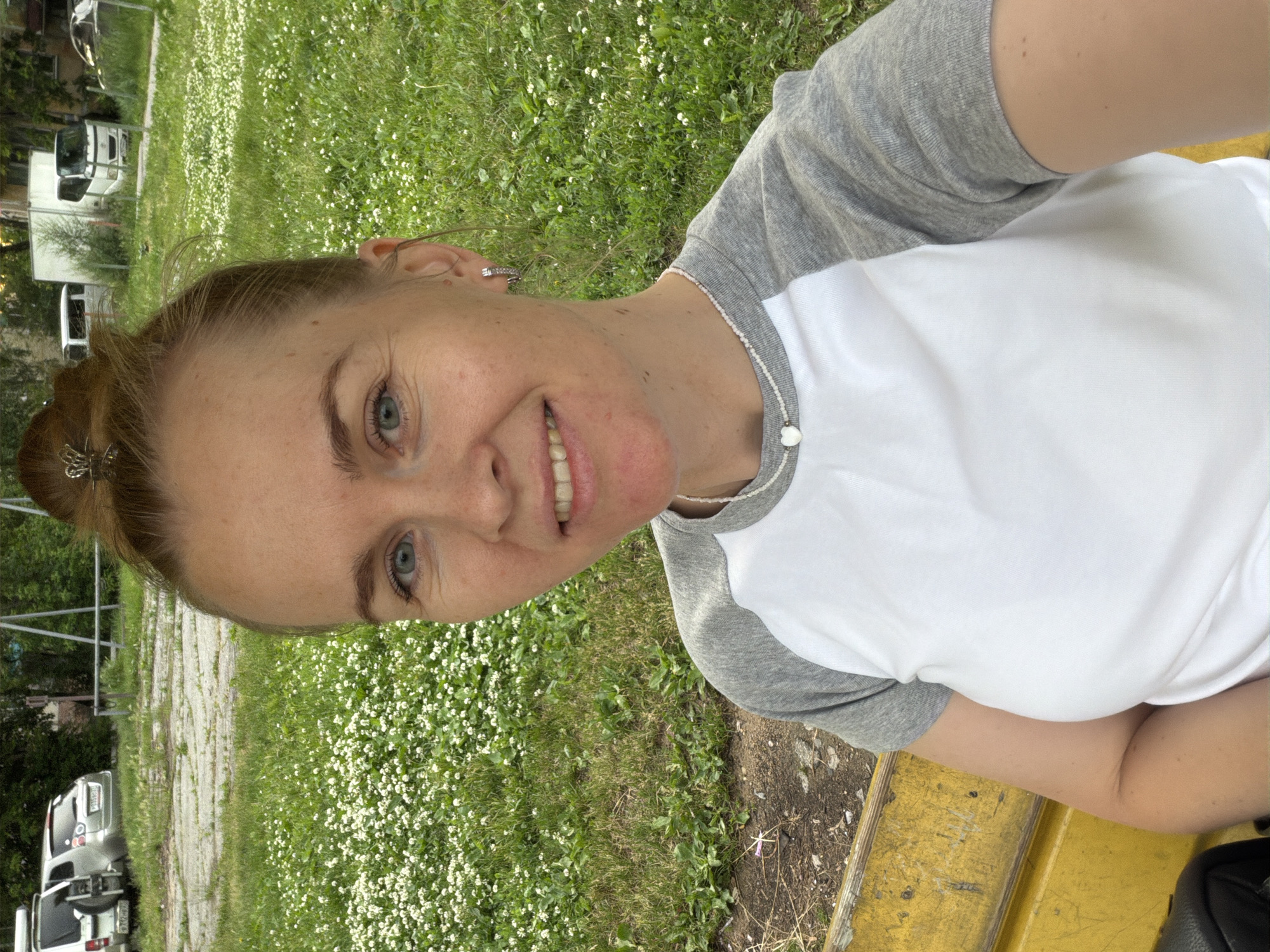 A young woman with blonde hair in a bun is taking a selfie in a park-like setting. She is wearing a white and grey raglan t-shirt and a silver necklace with a heart-shaped pendant. She is smiling and looking at the camera. Behind her, there is a grassy area with small white flowers, a yellow bench, and in the background, several cars and buildings are visible. The scene appears to be in an urban environment, likely in Khabarovsk, Russia, given the presence of various vehicle models common in the region and the general landscape.