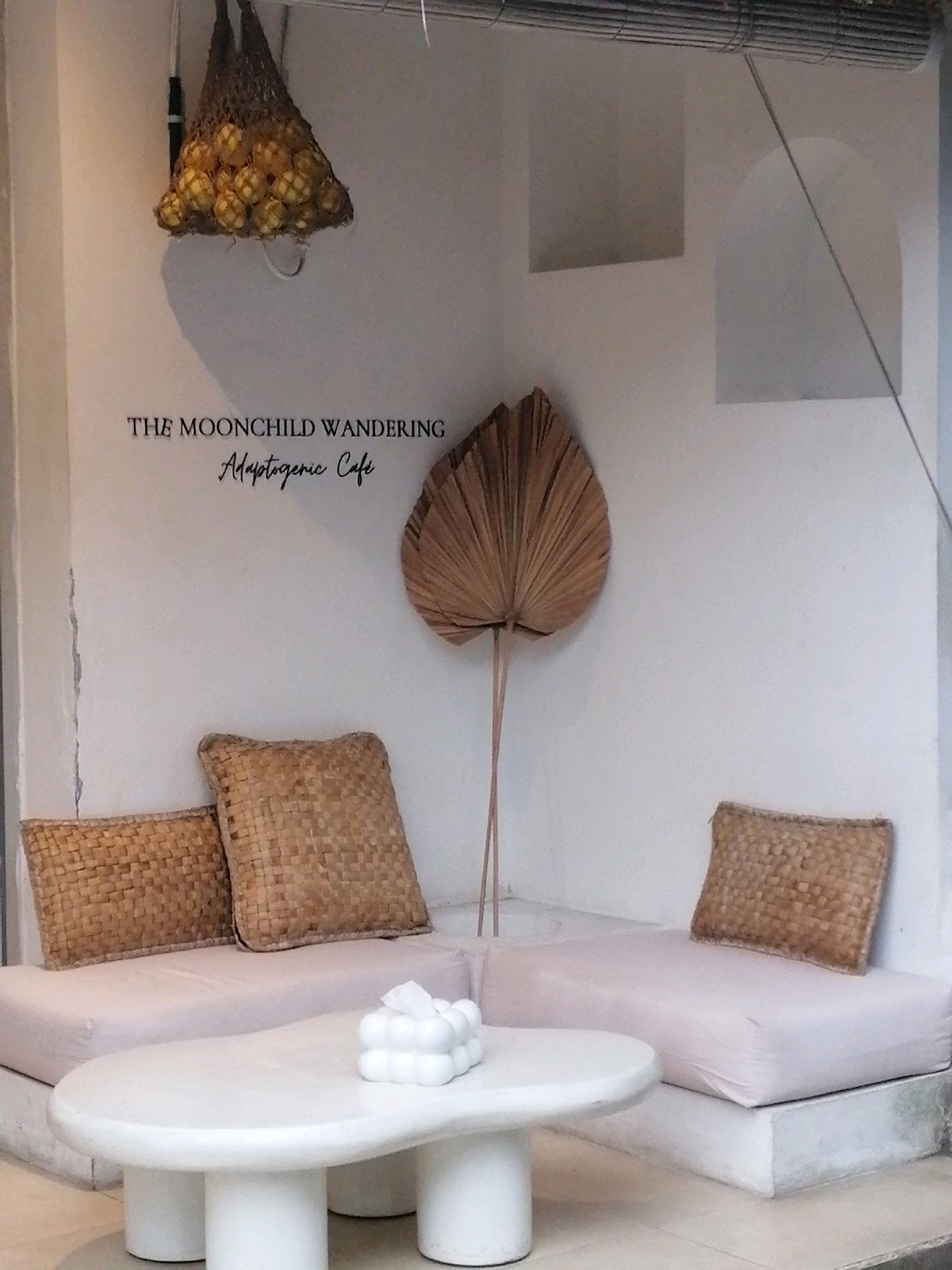 The image depicts an outdoor seating area at "The Moonchild Wandering Adaptogenic Cafe" in Ubud District, Indonesia. A white, organically shaped coffee table with three cylindrical legs sits in front of a light pink sectional sofa. Two woven square pillows rest on the left side of the sofa, and another woven pillow is on the right side. A fan-shaped dried palm leaf is mounted on the white wall behind the sofa. A net bag filled with yellow produce, possibly lemons or limes, hangs from the ceiling on the left. The wall features two recessed alcoves, one square and one arched. A contemporary white tissue holder with a bubble design sits on the coffee table.