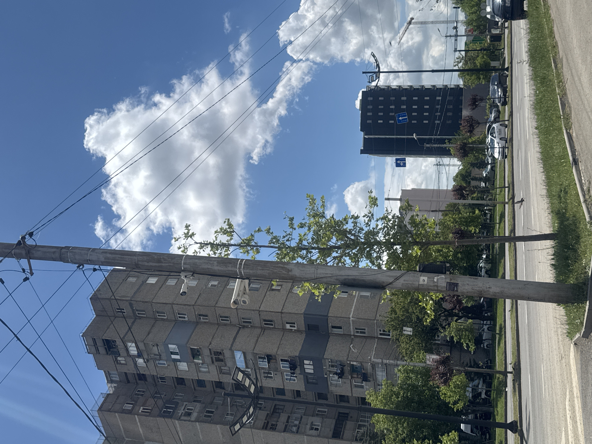 The image shows a street in Tbilisi, Georgia. On the left is a tall, gray, Soviet-era apartment building with many windows and at least one security camera mounted on the exterior. Utility poles and overhead wires are visible in the foreground. Across the street is another tall building, darker in color. Several cars are parked along the side of the road, and trees line the street. The sky is blue with white clouds. There are no people visible in the image.