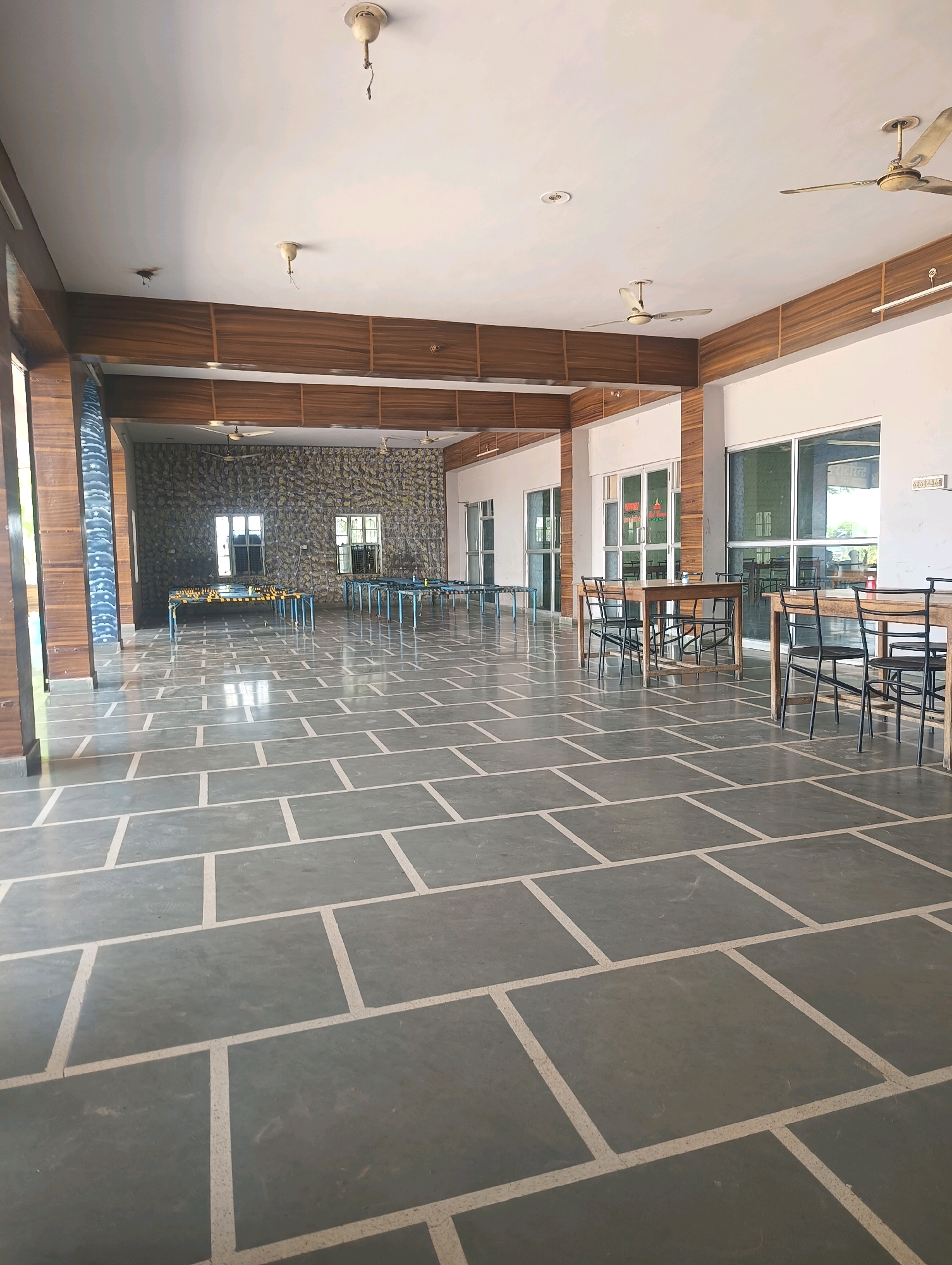 This image depicts an interior space, likely a large hall or common area, with a stone-tiled floor. The hall features wooden-paneled walls and a white ceiling with visible ceiling fans and light fixtures. In the background, there are several tables and chairs arranged, suggesting a dining or seating area. Large windows and glass doors are visible on the right side of the image, offering a glimpse of the outdoors. On the left side, a decorative pillar with a patterned design is prominent. The space appears to be unoccupied at the time the photograph was taken, and the overall impression is one of a functional, yet somewhat empty, establishment in Chhapri Bari, India.