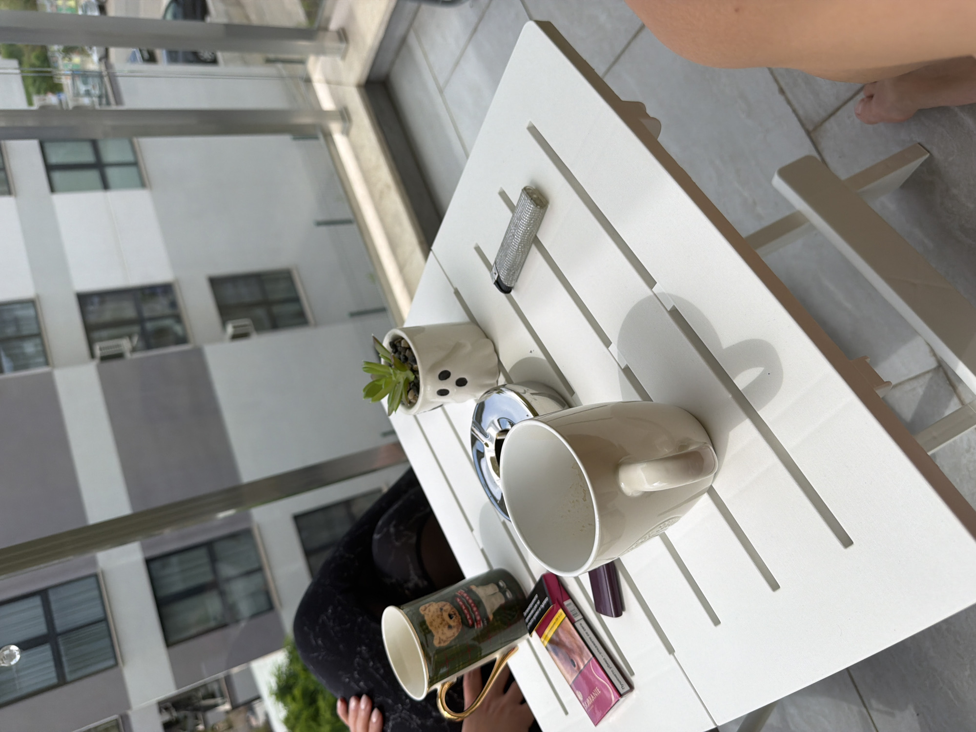 The image depicts a close-up of a white slatted outdoor table on a tiled balcony. The table holds two mugs, one dark green with a teddy bear design and a gold handle, and another plain beige mug. Also on the table are a silver ashtray, a pack of cigarettes, a lighter, and a small potted succulent in a white planter shaped like a ghost. In the background, a modern apartment building with gray and white horizontal stripes is visible, suggesting an urban setting. The balcony appears to overlook a street with parked cars. The perspective suggests the photo was taken from a seated position on the balcony.