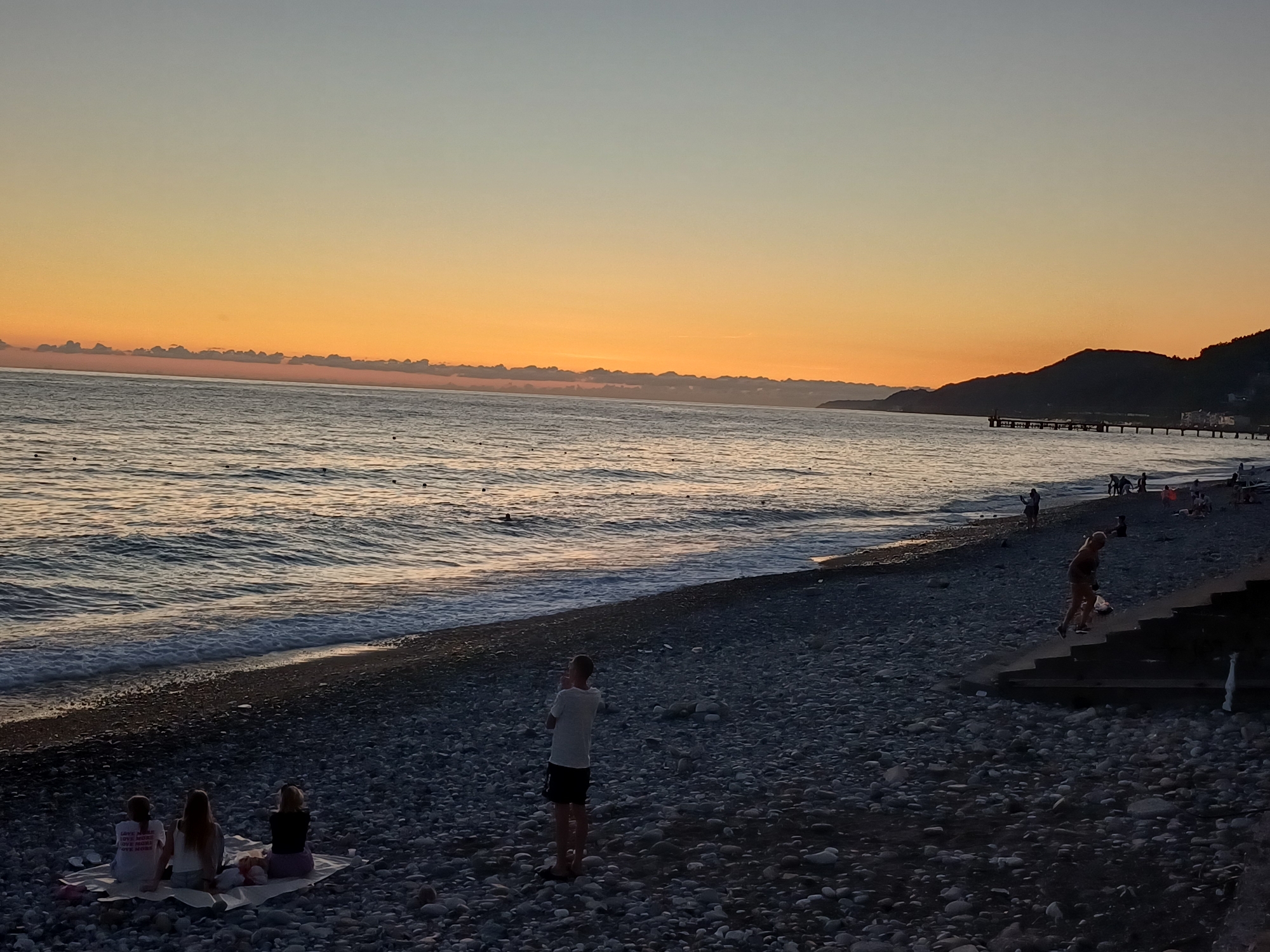 The image depicts a tranquil scene at sunset on a pebble beach in Sochi, Russia. The sky displays a gradient of warm colors, transitioning from a pale blue at the top to a vibrant orange and yellow near the horizon, with a line of clouds accentuating the horizon. The sea, with gentle waves lapping at the shore, reflects the warm hues of the sky. In the foreground, several people are scattered across the beach. Three individuals are seated on a blanket, facing the sea, seemingly enjoying the view. Further down the beach, a person in a white t-shirt and dark shorts stands looking out at the water. On the right side of the image, a set of stairs leads down to the beach, with a person walking down them. In the middle ground, a pier extends into the sea, and several more individuals are visible on the beach and near the water. The overall atmosphere is peaceful, with the setting sun casting a soft glow over the landscape.