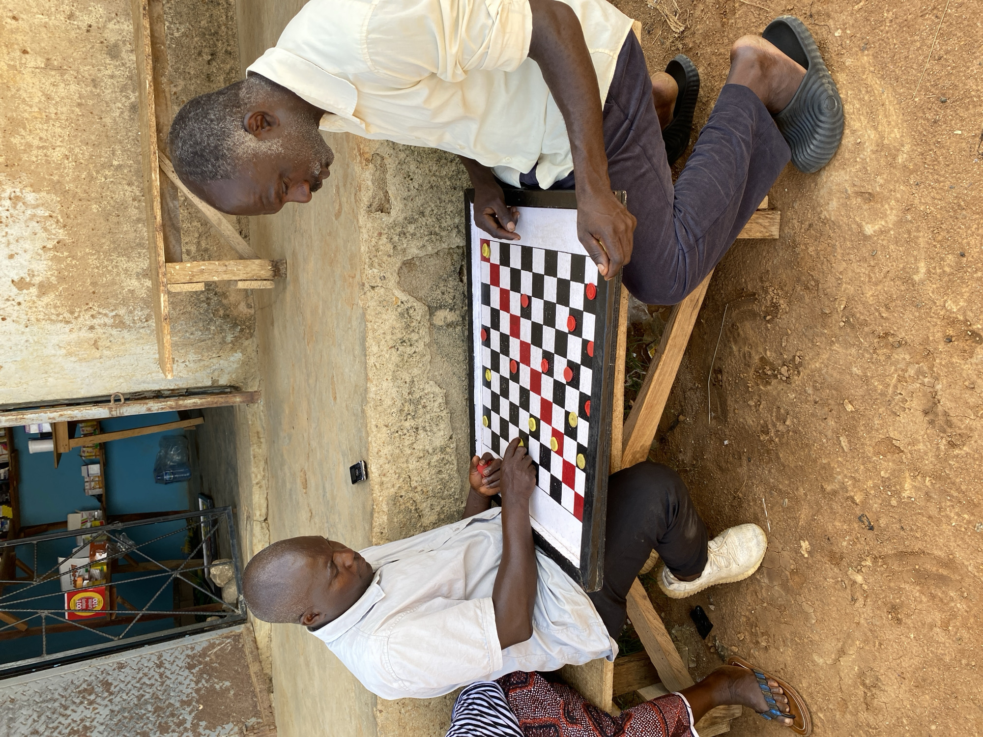Two African men are playing draughts or checkers outside a building in what is presumed to be Riyom, Nigeria. One man wears a light-colored shirt and dark pants and sits on a wooden frame. He is wearing dark slides. The other man wears a similar shirt and patterned trousers, with one white shoe and one sandal. They are both focused on a draughts board, which is vertically oriented between them. The area surrounding the men is a dirt or stone surface. Behind one man is a storefront, or shop, visible through a metal grate. Behind the other is a concrete wall.