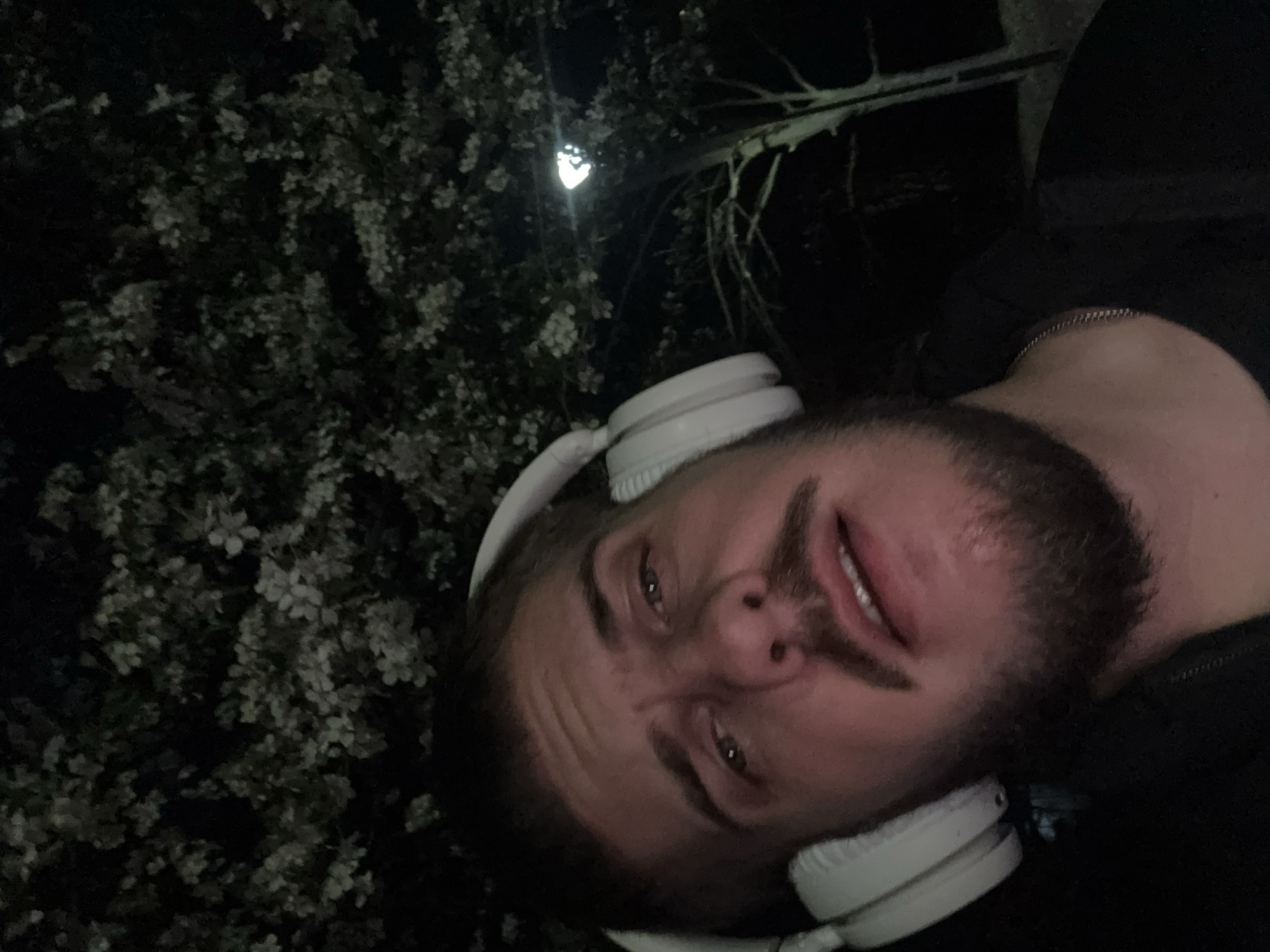 A man with a beard and white headphones lies under a flowering tree at night, potentially in Sofia, Bulgaria, given the prompt. He is positioned on his side, facing the camera. The scene is illuminated by a light source above and behind the tree, which is covered in white blossoms. He wears a dark-colored garment. A chain is visible around his neck.