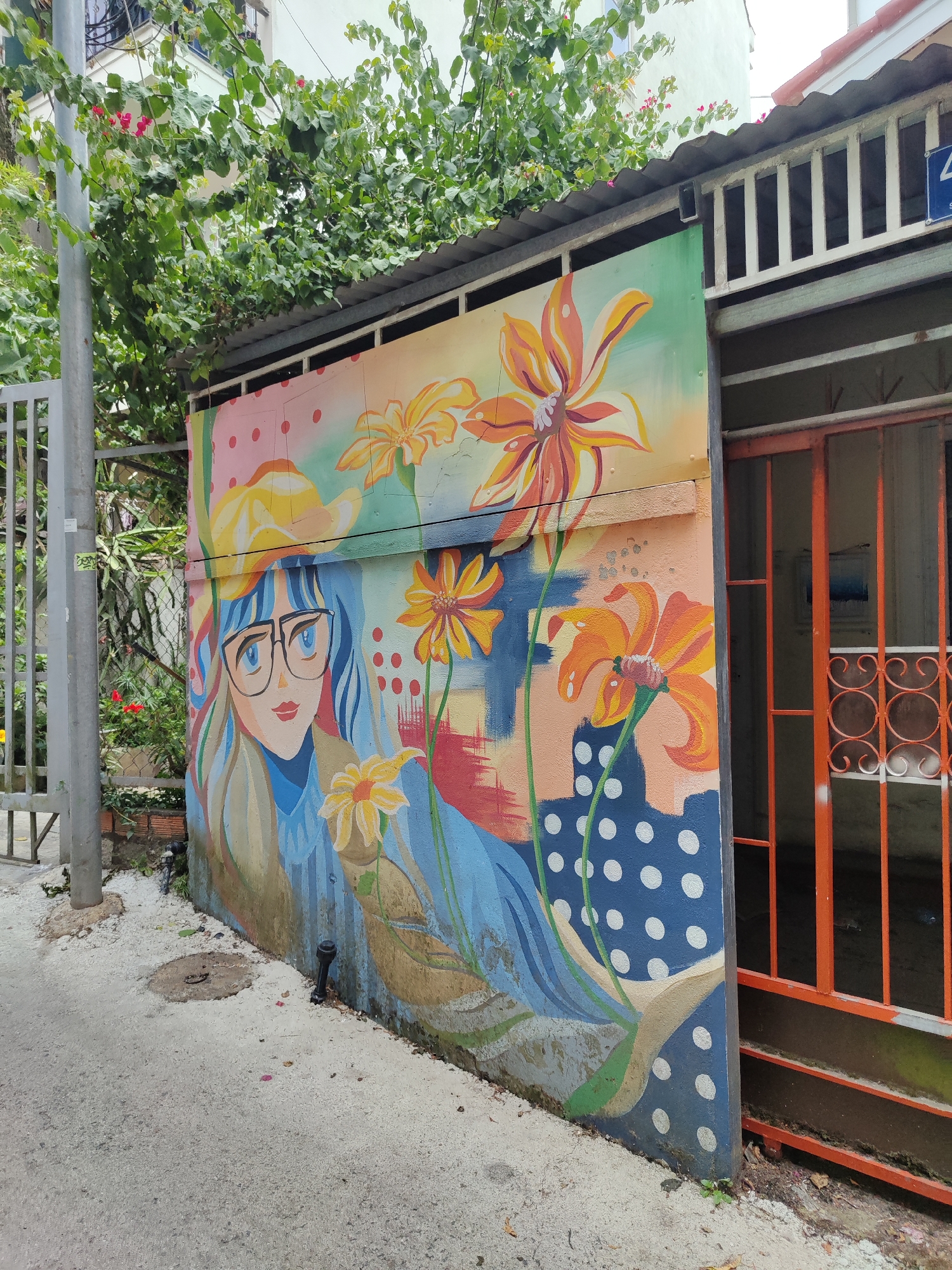 A vibrant mural adorns a wall in Da Lat, Vietnam. The artwork depicts a woman with blue hair, glasses, and a yellow hat, surrounded by stylized orange and yellow flowers. A building with a blue and white polka-dot pattern is incorporated into the design. The mural is painted on a concrete wall next to an orange metal gate and a grey post. Lush greenery overhangs the wall. The ground is concrete with scattered debris.