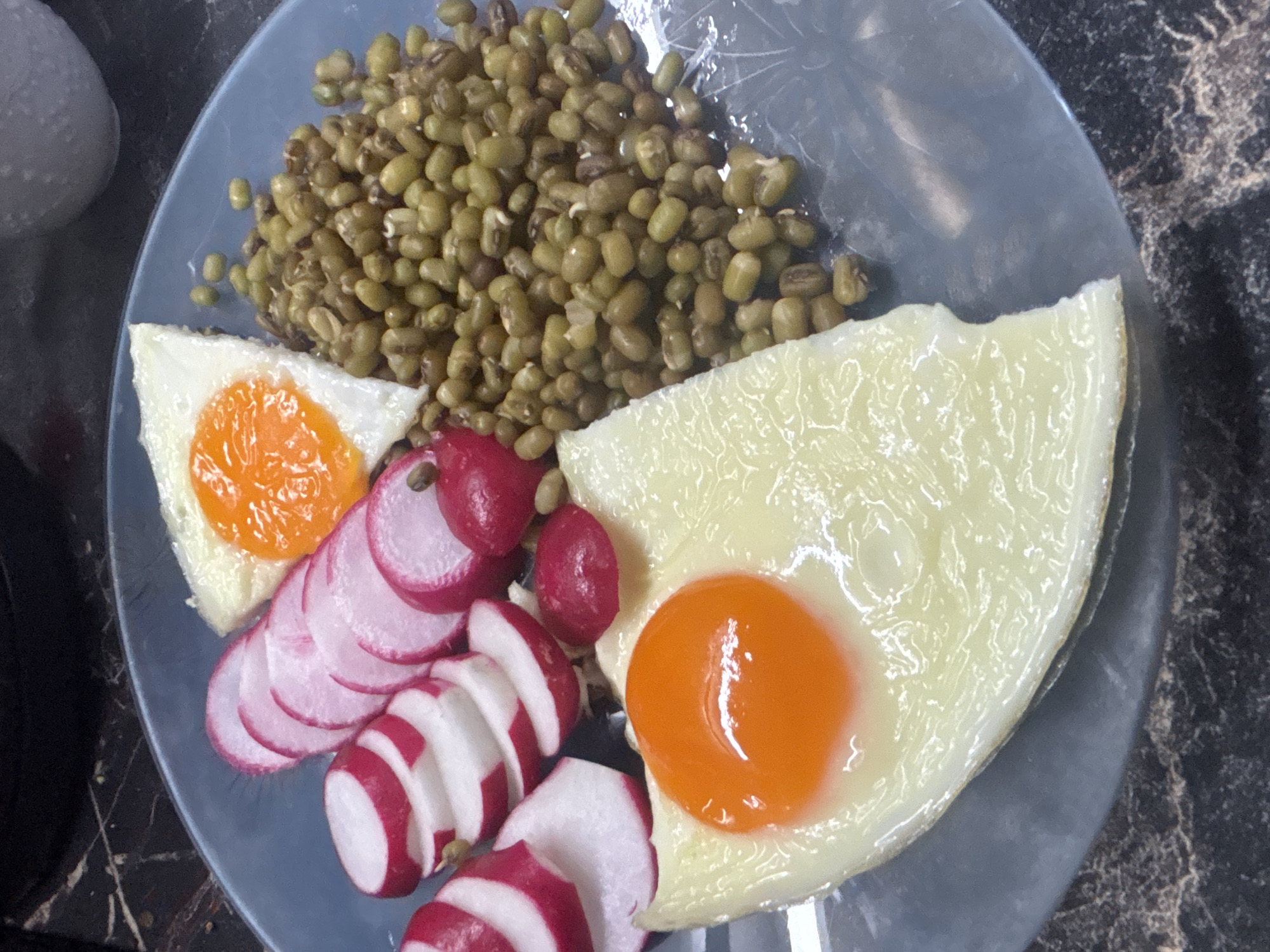 The image shows a plate of food on a dark marble surface. The plate contains two fried eggs, one with a runny yolk and the other with a set yolk. There are also sliced radishes, a pile of cooked mung beans, and a small, dark object, possibly a piece of food or a garnish. The lighting suggests an indoor setting. There is no visual information in the image that would allow for the determination of its location in the city of Sochi, Russia.