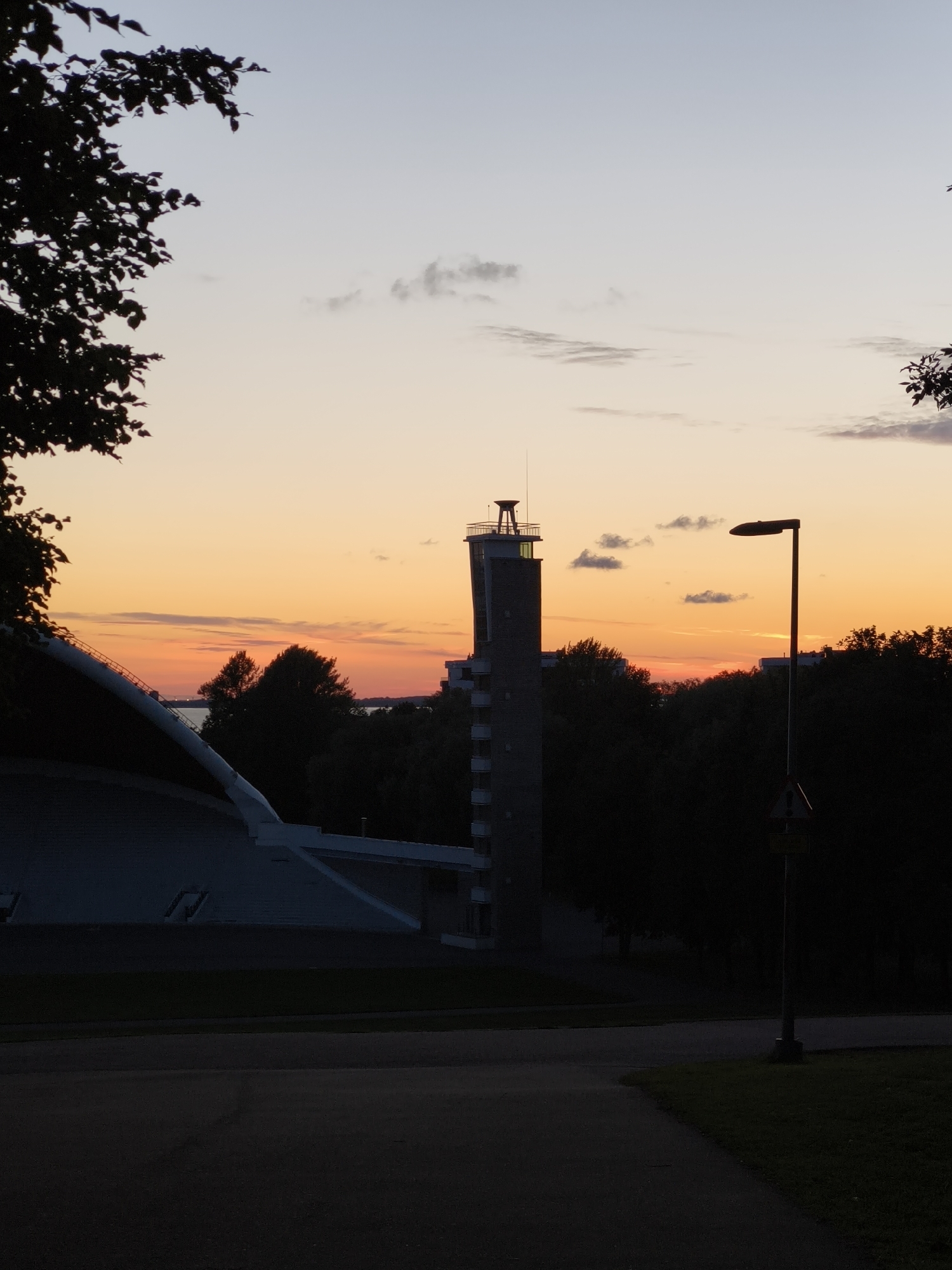 This image captures a sunset scene in Tallinn, Estonia, likely near the Pirita district, which is known for its seaside location and architectural landmarks. In the foreground, a dark silhouette of a paved pathway leads the eye towards the middle ground. To the left, a portion of a large, modern structure with a curved, white roofline dominates the frame, suggesting an amphitheater or stadium.
The middle ground is filled with dark silhouettes of trees, with a tall, slender tower, possibly a fire watchtower or part of a communications building, rising above them on the right side of the tower is a building with many windows. The tower is constructed of brick and has a distinctive top section with lights. A tall, dark lamppost stands in front of the tower, its silhouette stark against the colorful sky. A traffic sign is visible at the base of the lamppost.
The background is a vibrant sunset sky, transitioning from pale blue at the top to warm oranges and pinks near the horizon. Wispy clouds are scattered across the sky, catching the light of the setting sun. A body of water, likely the Baltic Sea, is visible in the distance behind the trees. The overall impression is one of quiet beauty, with the silhouetted urban and natural elements creating a dramatic contrast with the colorful sky.