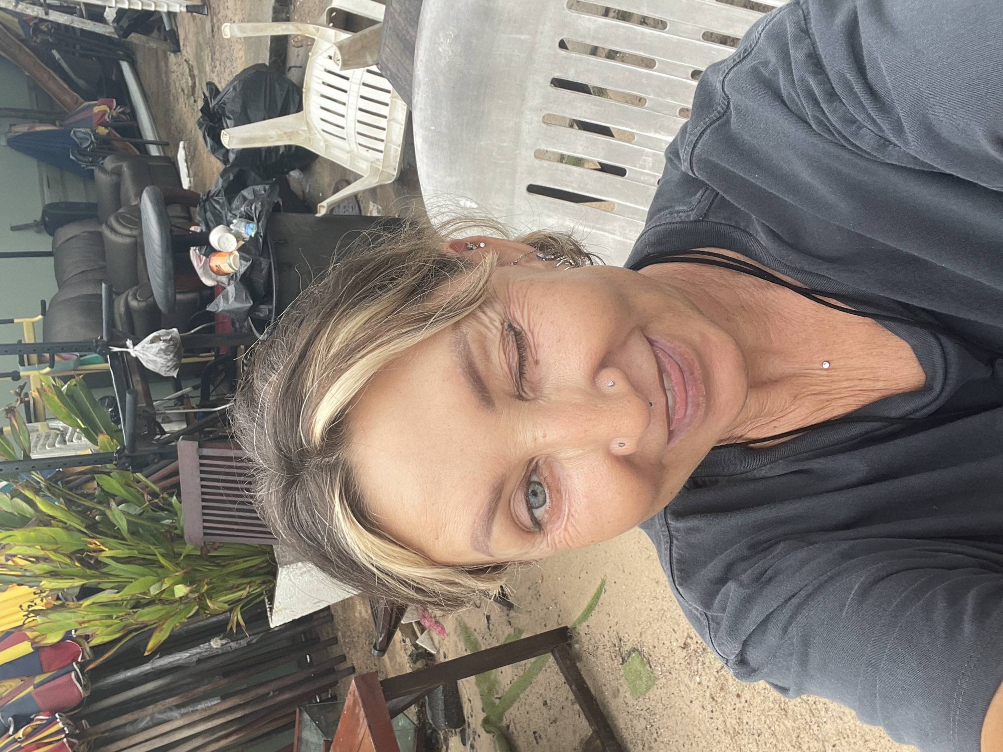 The image is a selfie of a light-skinned woman with graying blonde hair, a black t-shirt, and multiple piercings in her ear, nose, and neck. She is winking. The background appears to be an outdoor cluttered space, with discarded tires, black garbage bags, plastic chairs, and what may be construction materials. There are also some green leafy plants visible. The location appears to be in Choeng Thale, Thailand.