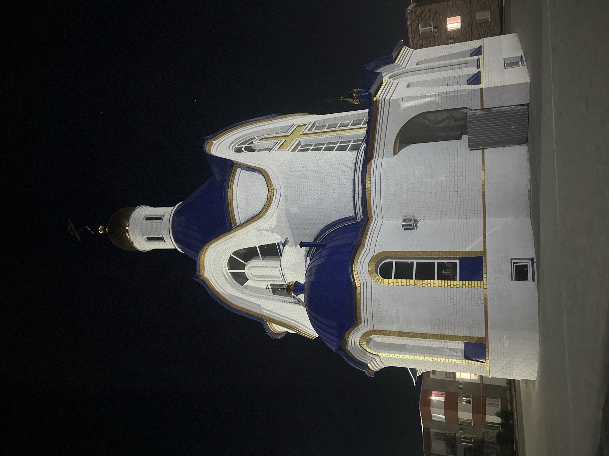 A low-angle, full exterior shot captures a church at night in Nezhinka, Russia. The church is primarily white brick with blue domes and gold trim. The main structure has a large central dome topped with a cross. Smaller blue domes are visible on the sides. The building features arched windows with a gold cross design on one large window. To the left of the church is a multi-story residential building with orange and white facades and illuminated windows. To the right, another brick building with lit windows is partially visible. The ground in front of the church is paved. The sky above is dark, with a few faint stars visible.