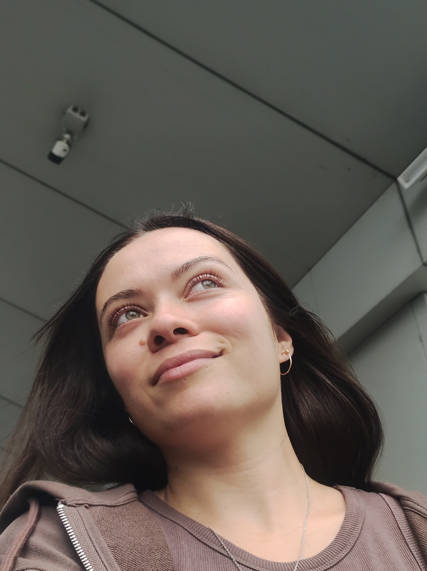 The image is a close-up, low-angle shot of a young woman looking upwards and to her right. She has long, dark brown hair and light-colored eyes. She is wearing a brown hooded sweatshirt with a zipper and a silver necklace. A small gold hoop earring is visible in her right ear. In the background, a security camera is mounted on a gray ceiling with angled structural elements visible to the right. The setting appears to be an indoor or sheltered urban environment. The image does not provide any specific visual cues to determine its location within the city of Kirov, Russia.