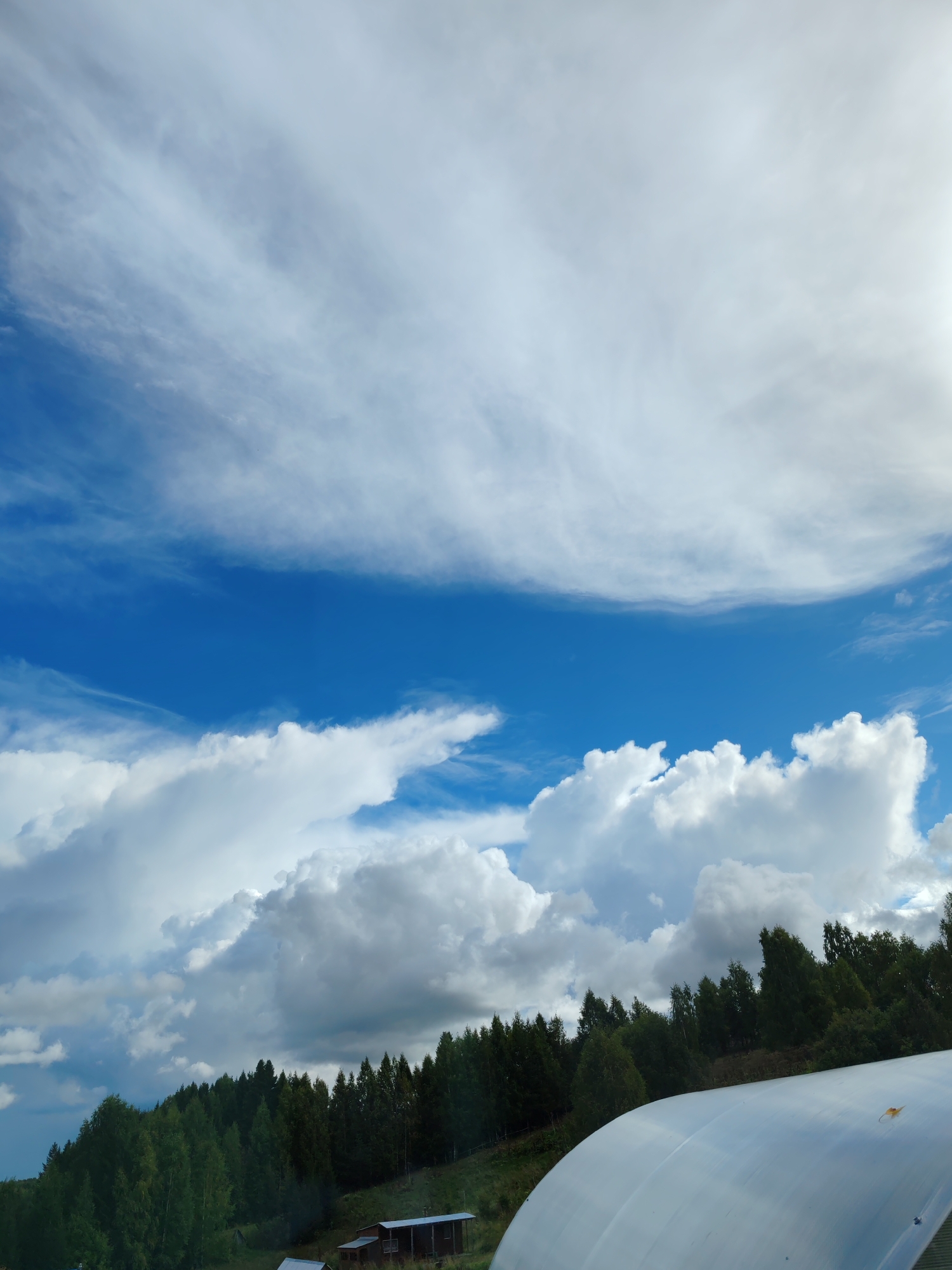 The image depicts a rural landscape under a bright, cloudy sky. The foreground is dominated by a large, white, curved structure, possibly a greenhouse or a temporary shelter, partially obscuring the view. Beyond this structure, a dense line of coniferous trees forms a dark green barrier along a sloping hillside. Below the tree line, a small, rustic building with a dark roof is visible. The sky is a vibrant blue, layered with various types of clouds, including wispy cirrus formations high up and larger, cumulus clouds lower down, suggesting dynamic weather conditions. The sun is partially visible, casting a bright light on the scene. The location is described as Чухлэм, Russia.