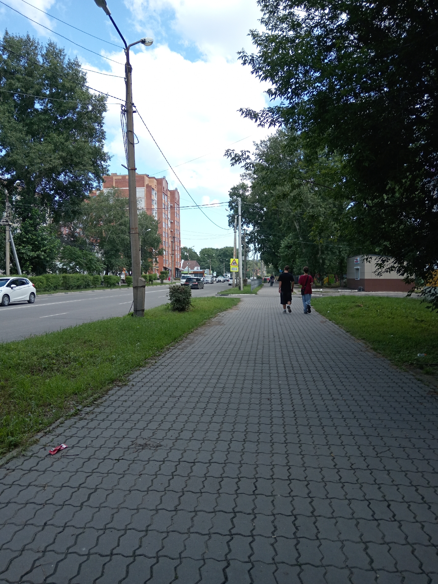 A wide shot captures a paved sidewalk and an adjacent road in Belogorsk, Russia, under a partly cloudy sky. Two individuals, one wearing a black t-shirt and shorts, and the other a red t-shirt and jeans, walk away from the camera on the sidewalk. The sidewalk is made of interlocking concrete pavers. To the left of the sidewalk, a grassy area borders a road with several cars. Further left, a multi-story brick apartment building is visible, with trees lining the street. A lamppost stands near the edge of the sidewalk, with visible wires. On the right side of the sidewalk, trees with dense foliage line the path, and a small, low building with a brown and beige facade is present. In the foreground of the sidewalk, a crumpled red wrapper lies on the ground.