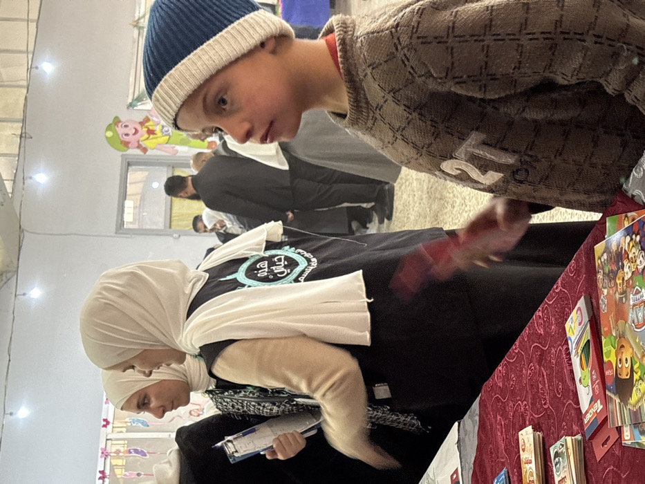 This image depicts an indoor scene in Kafr Batna, Syria, at what appears to be a community event or exhibition. In the foreground, a young boy is visible from the chest up, wearing a blue and white knit beanie and a brown patterned sweater with "2T" embroidered in white. He faces right in profile, looking upwards and right with a neutral expression.
To the boy's left, a table is covered with a red, intricately patterned tablecloth. On the table are several stacks of Syrian Pound banknotes, children's activity books (including one featuring "Paw Patrol"), and a box of "Tender" colored pencils.
Behind the table, two women are partially visible. The woman on the left wears a light-colored hijab and a black and white patterned top, holding a blue clipboard with papers; her right arm is slightly blurred, indicating motion. The woman to her right wears a white hijab and a black vest adorned with a light blue circular logo featuring Arabic text, specifically "هذه حياتي" (This is my life). Her left arm has a light beige sleeve, and her hand is blurred while holding a red object.
In the midground and background, multiple adults are present, some in dark suits, others in traditional attire including hijabs. A large cartoon cutout of a girl in a green hat and yellow top, holding a camera, is affixed to a wall in the background near a doorway. The indoor space has light-colored walls, a light-toned floor, and is illuminated by several bare light bulbs hanging from the ceiling. Colorful paper cutouts decorate a wall in the upper left background.