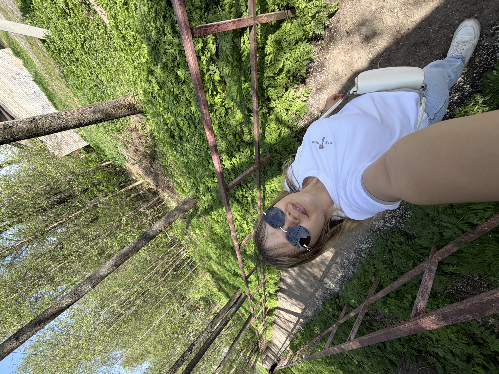 A selfie taken from a slightly elevated position along a tree-lined path or walkway. The photograph features a fair-skinned person, possibly female, wearing sunglasses and a white t-shirt. They are wearing light blue jeans and white sneakers. They are carrying a white shoulder bag. Rusty metal railing is visible in the foreground and background, suggesting a bridge or walkway structure. Lush green foliage and tall trees line the sides of the path, indicating a park or wooded area. The location may be in the city of Chaikovsky, Russia.