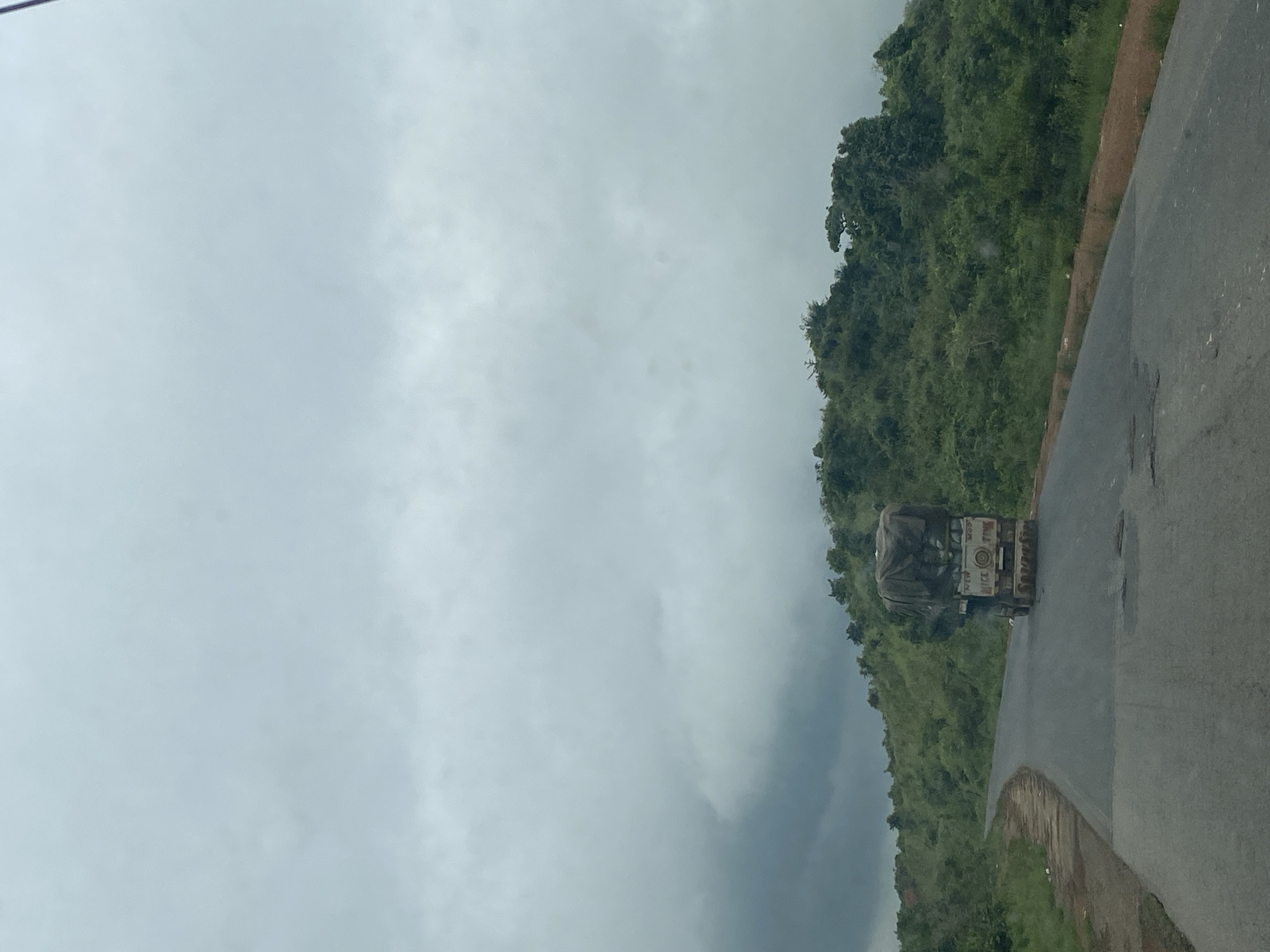 An overcast sky looms above a rural highway in Assob Hausa, Nigeria. A truck, its cargo covered by a tarp, travels on the road. Lush green vegetation covers the hillsides flanking the highway. The road itself shows signs of wear, with visible potholes. The scene appears to be captured from within a moving vehicle, given the perspective and the slight motion blur.