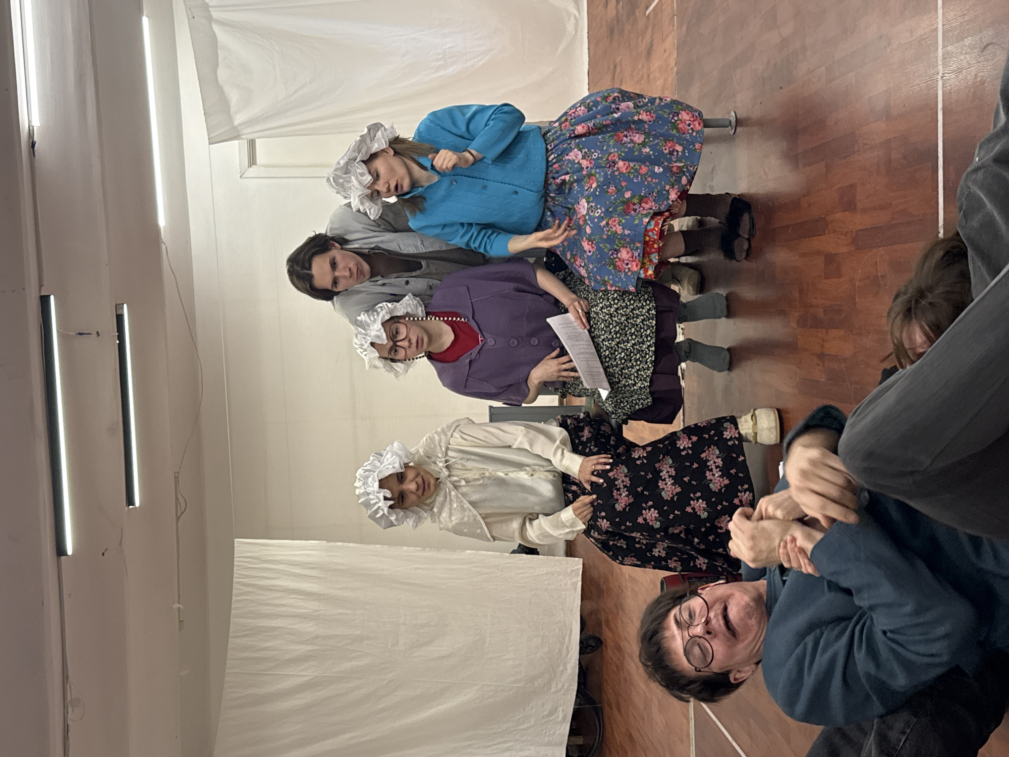 The image shows a group of people, presumably actors, in a room with a wooden floor. They are dressed in old-fashioned costumes, including bonnets and floral print skirts, suggesting they might be rehearsing or performing a play. One person is holding a script. Fluorescent lights are visible on the walls. It is unlikely this image is in Moscow, Russia.