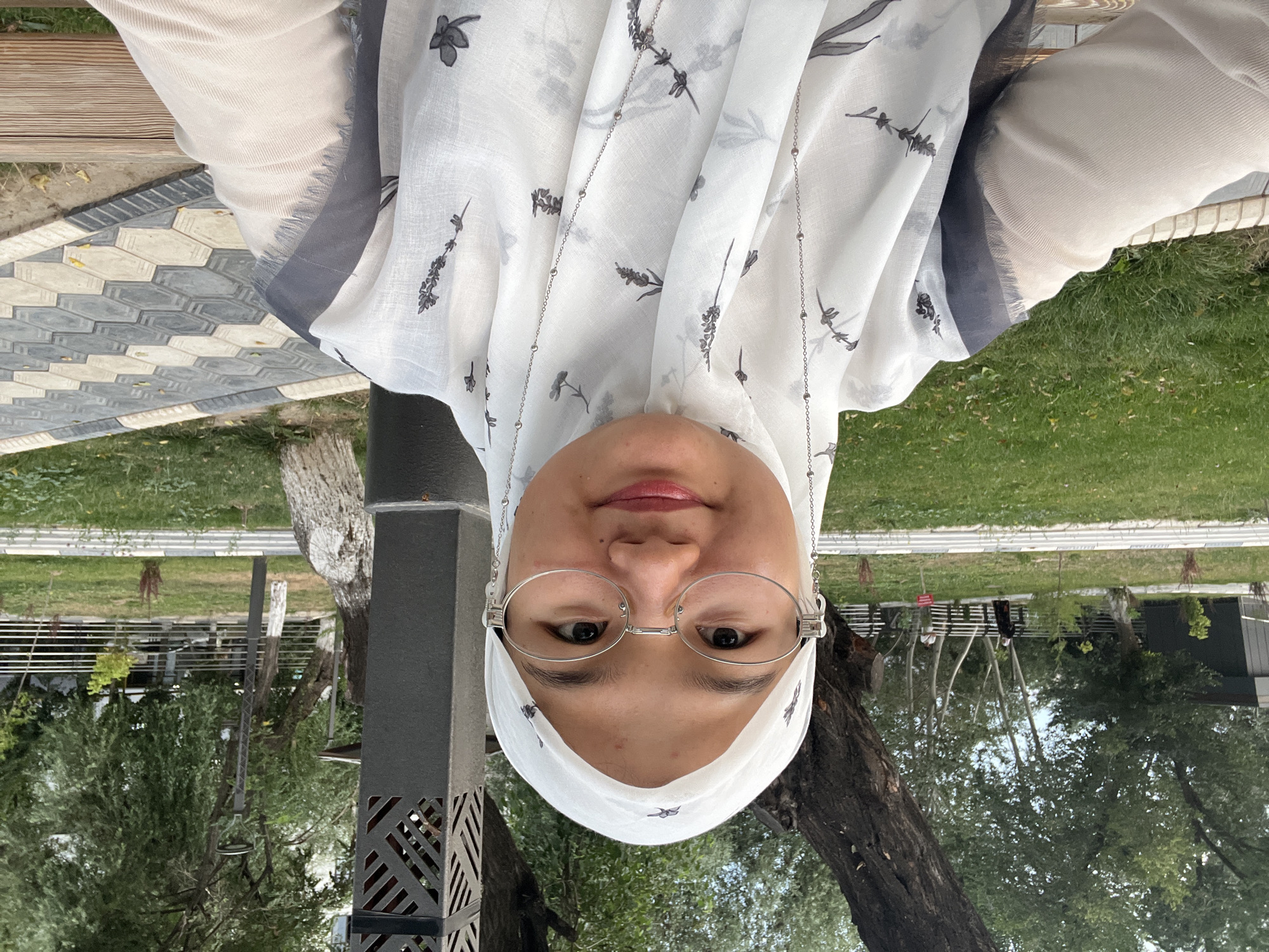 In Osh City, Kyrgyzstan, a young woman wearing a white hijab and a light-colored, patterned scarf is seated outdoors on a wooden bench. She is wearing glasses with silver frames. Behind her, a large tree trunk is visible, along with a black metal structure and a lamppost. The background includes a grassy area with scattered leaves, a paved walkway with hexagonal and diamond-shaped tiles, and more trees. Two other individuals are visible in the distance. The scene appears to be in a park or public outdoor space.