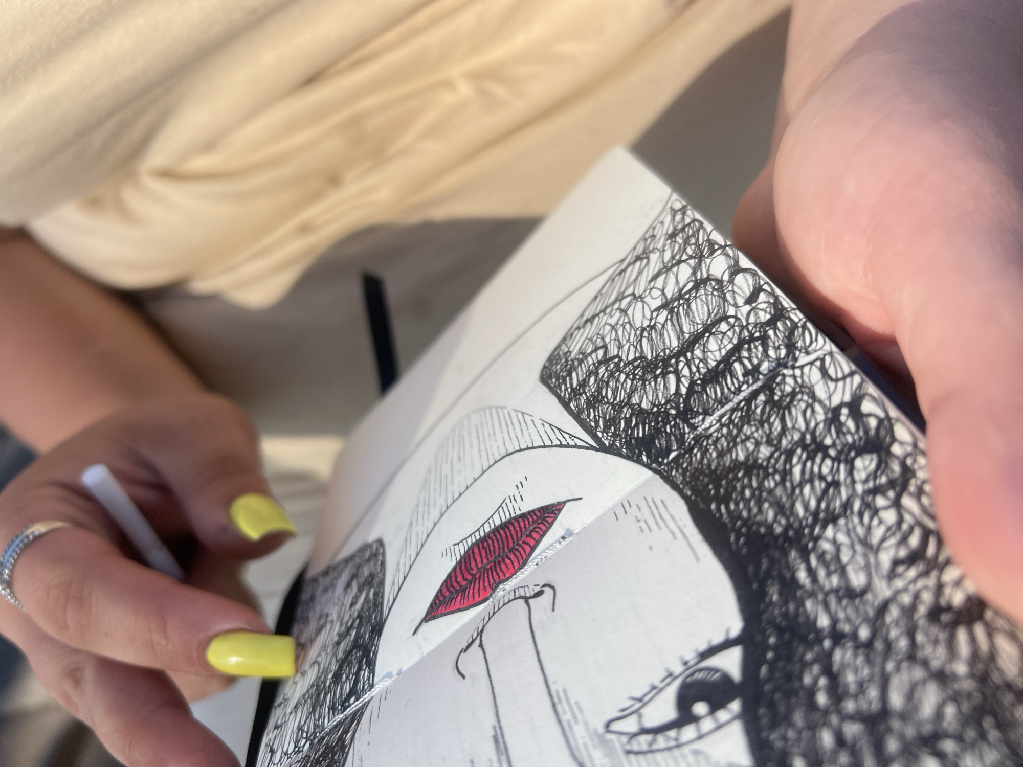 The image shows a close-up of a person's hands holding a sketchbook. The sketchbook contains a drawing of a face with detailed black ink lines and a red accent on the lips. The person's fingernails are painted bright yellow, and they are wearing a silver ring. One hand holds a white stylus or pen, poised as if to continue drawing. The background is blurred, showing a light-colored fabric, possibly clothing, and a hint of a shadow, suggesting an outdoor setting. The lighting appears to be natural, possibly indicating daytime. The focus is on the act of drawing and the partially completed artwork.